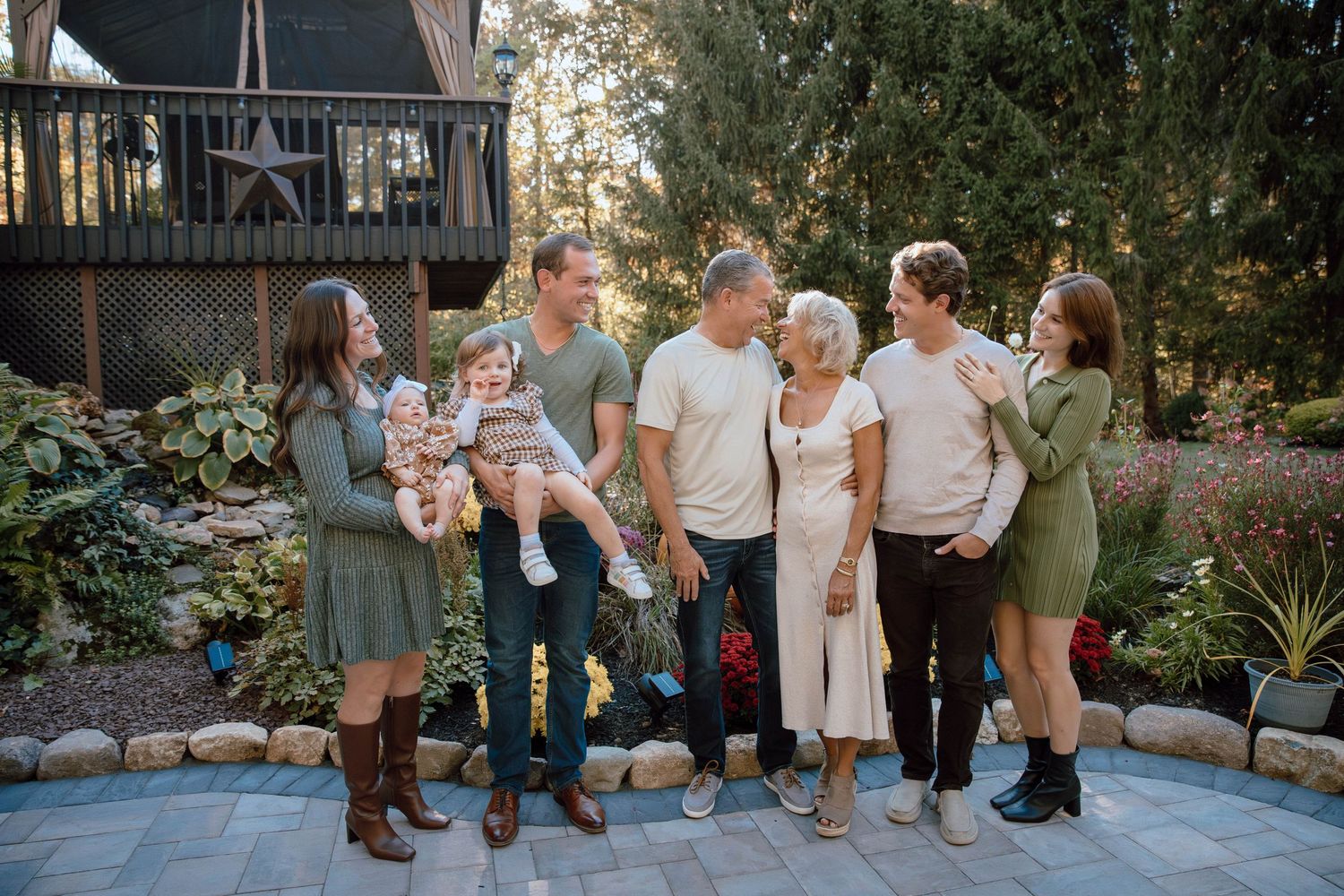 Multi-generational family of seven smiling together outdoors in a lush garden setting during a fall portrait session.