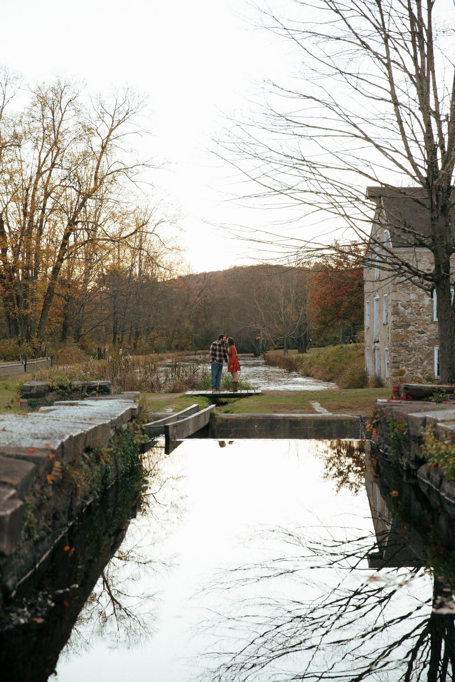 Couple standing on a canal lock bridge surrounded by autumn trees and calm water reflections in a historic park.