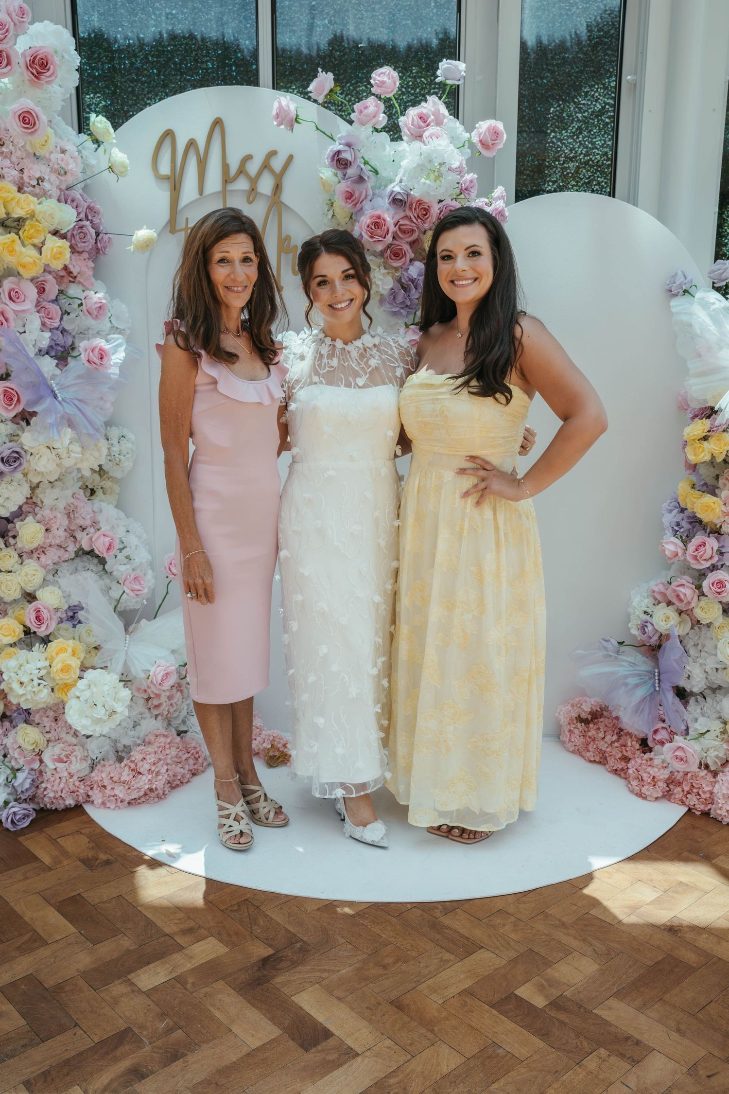 Bride in white floral gown poses with two guests in pink and yellow dresses before a floral arch.