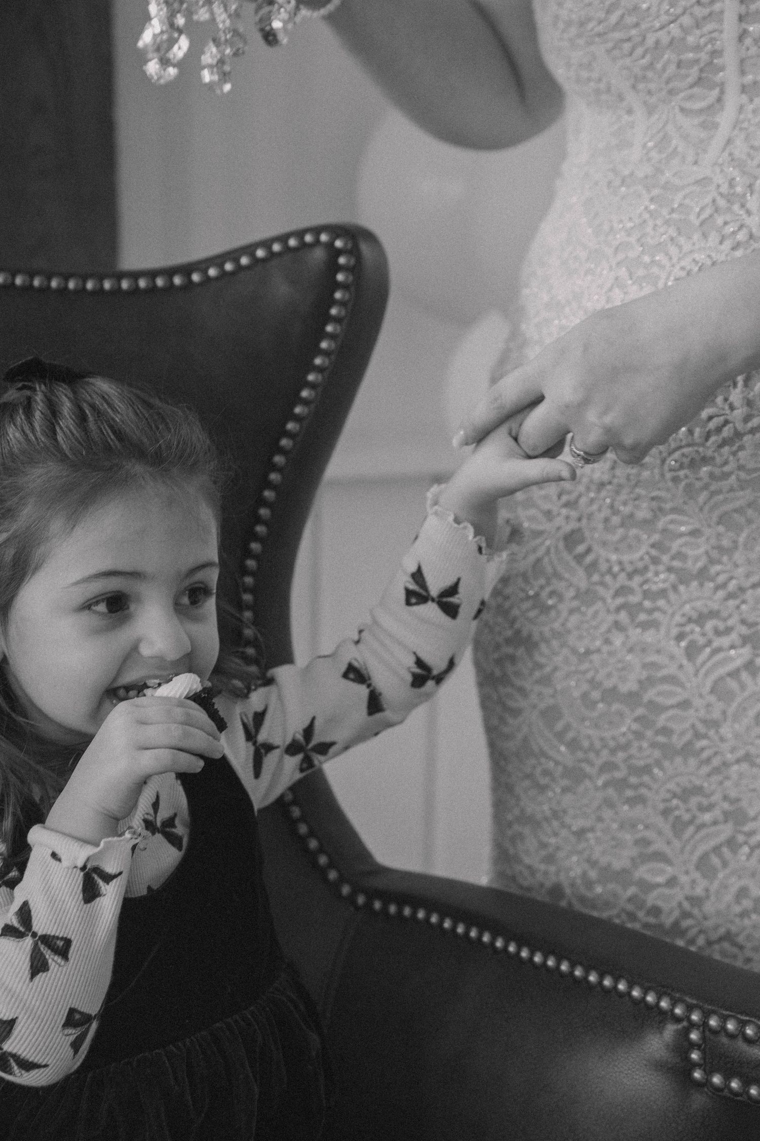 Black and white photo of a young girl eating a treat while holding hands with a woman in a lace dress near a leather chair.