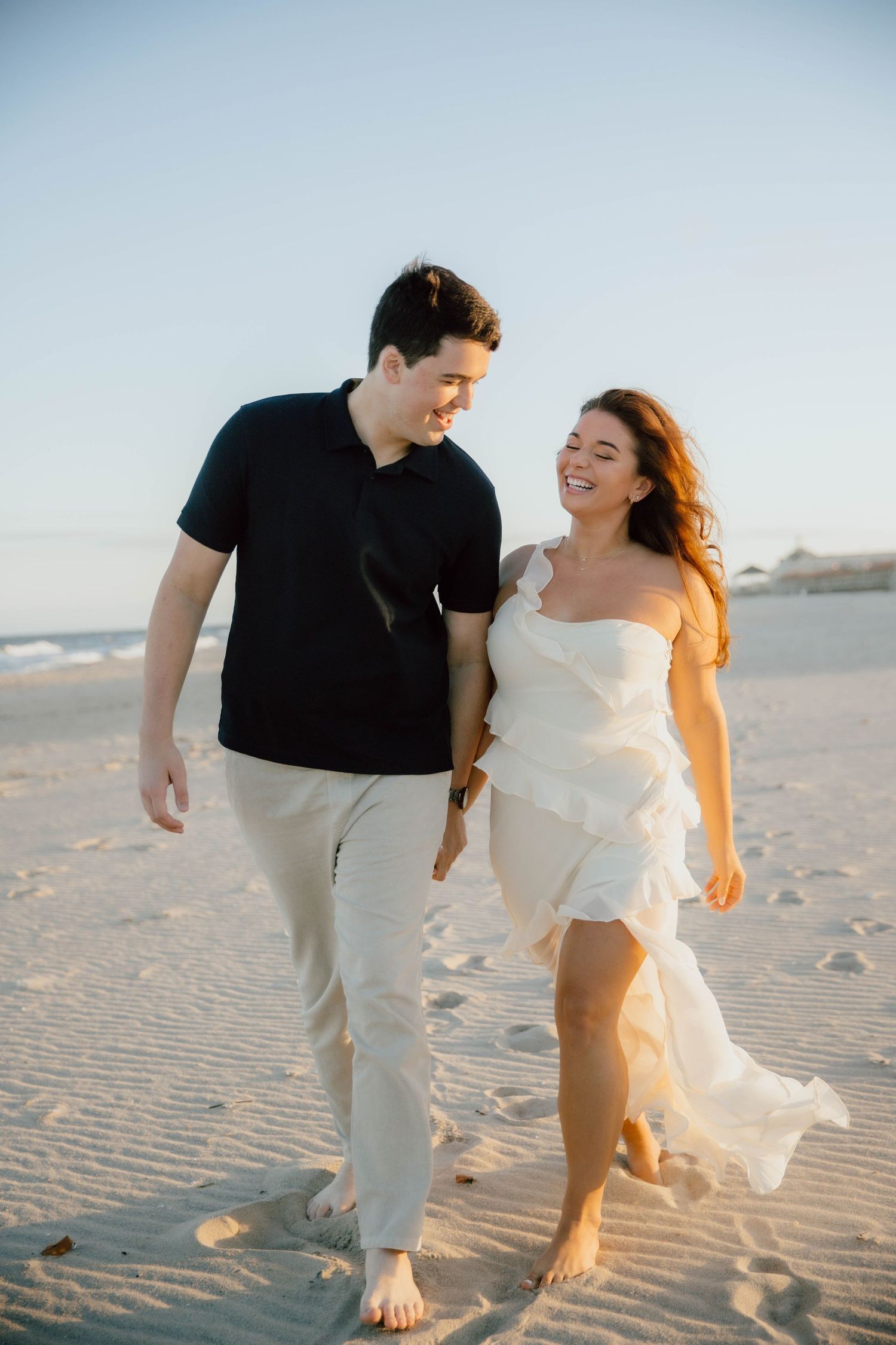 Smiling couple strolling barefoot on a sandy beach at golden hour, woman in white dress.