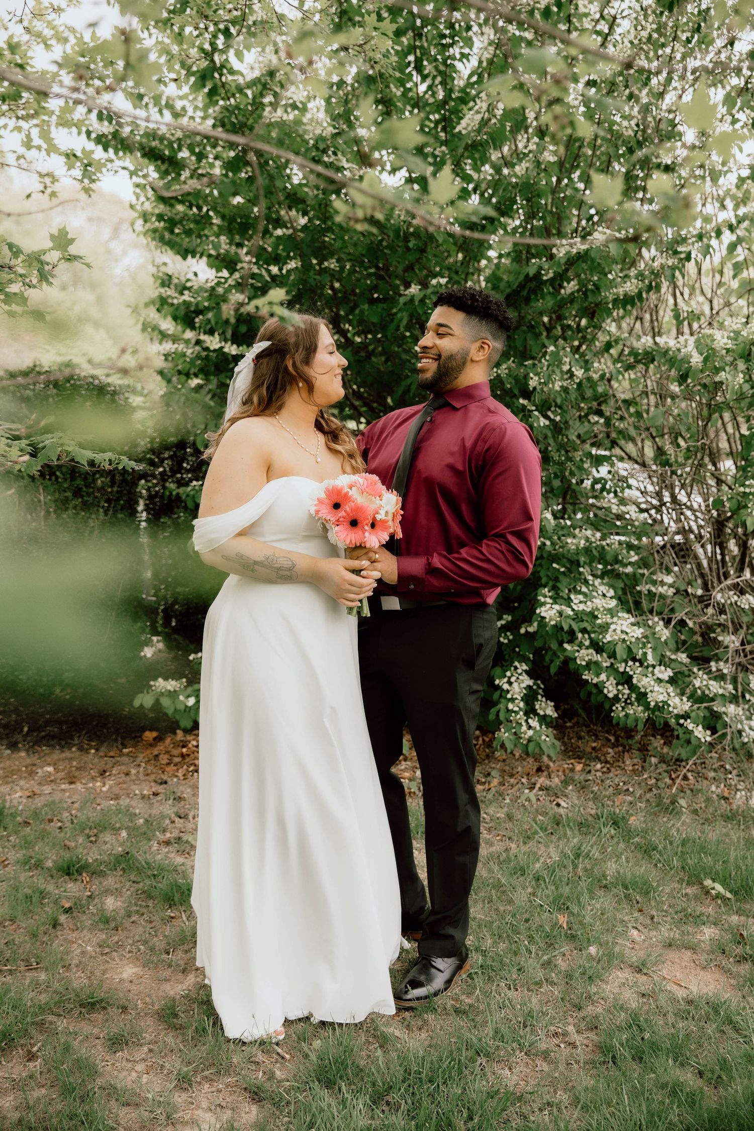 Bride in off-shoulder white gown and groom in burgundy shirt share loving look outdoors.
