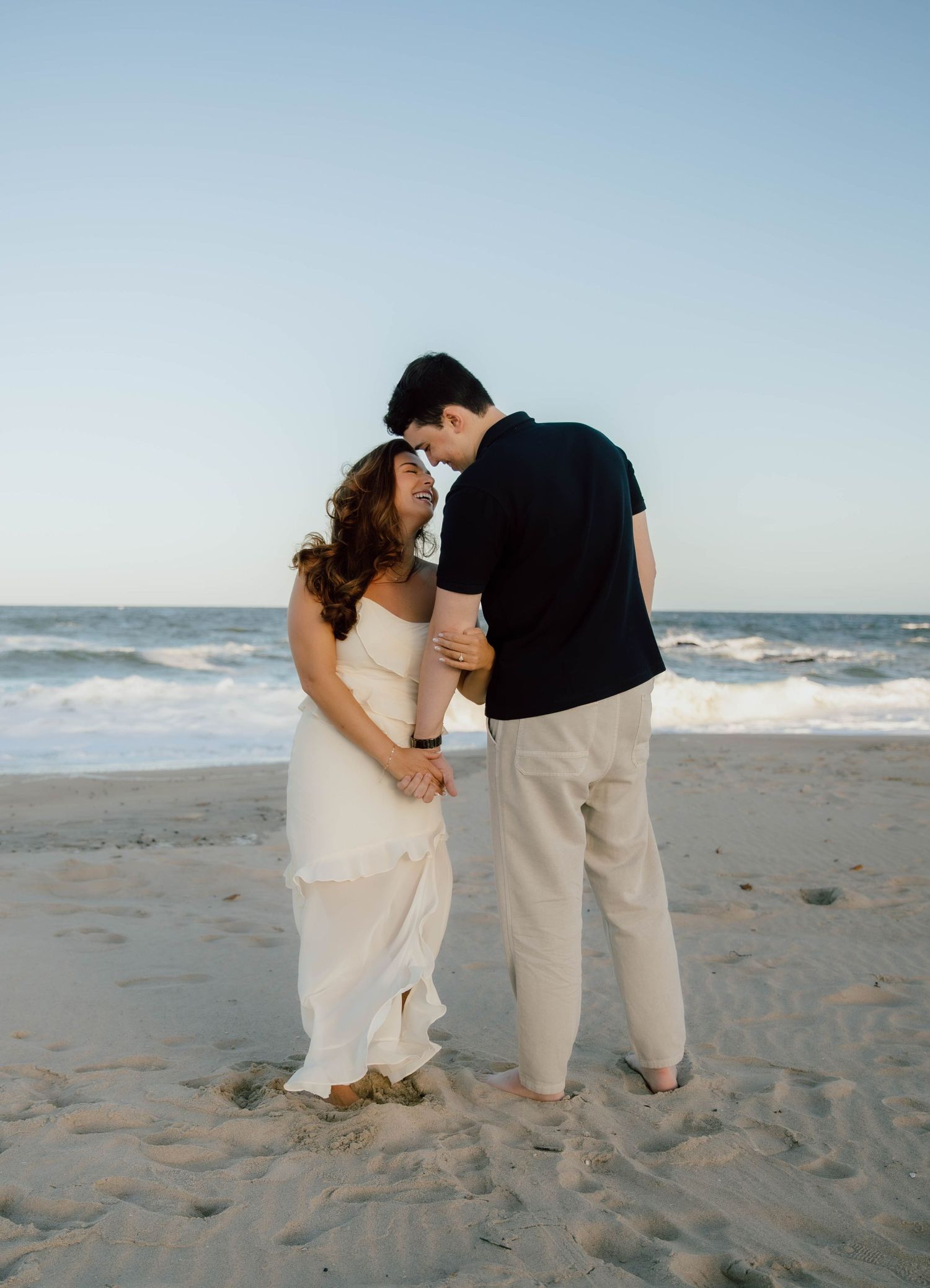 Romantic couple in white dress and black shirt embracing on sandy beach at dusk.