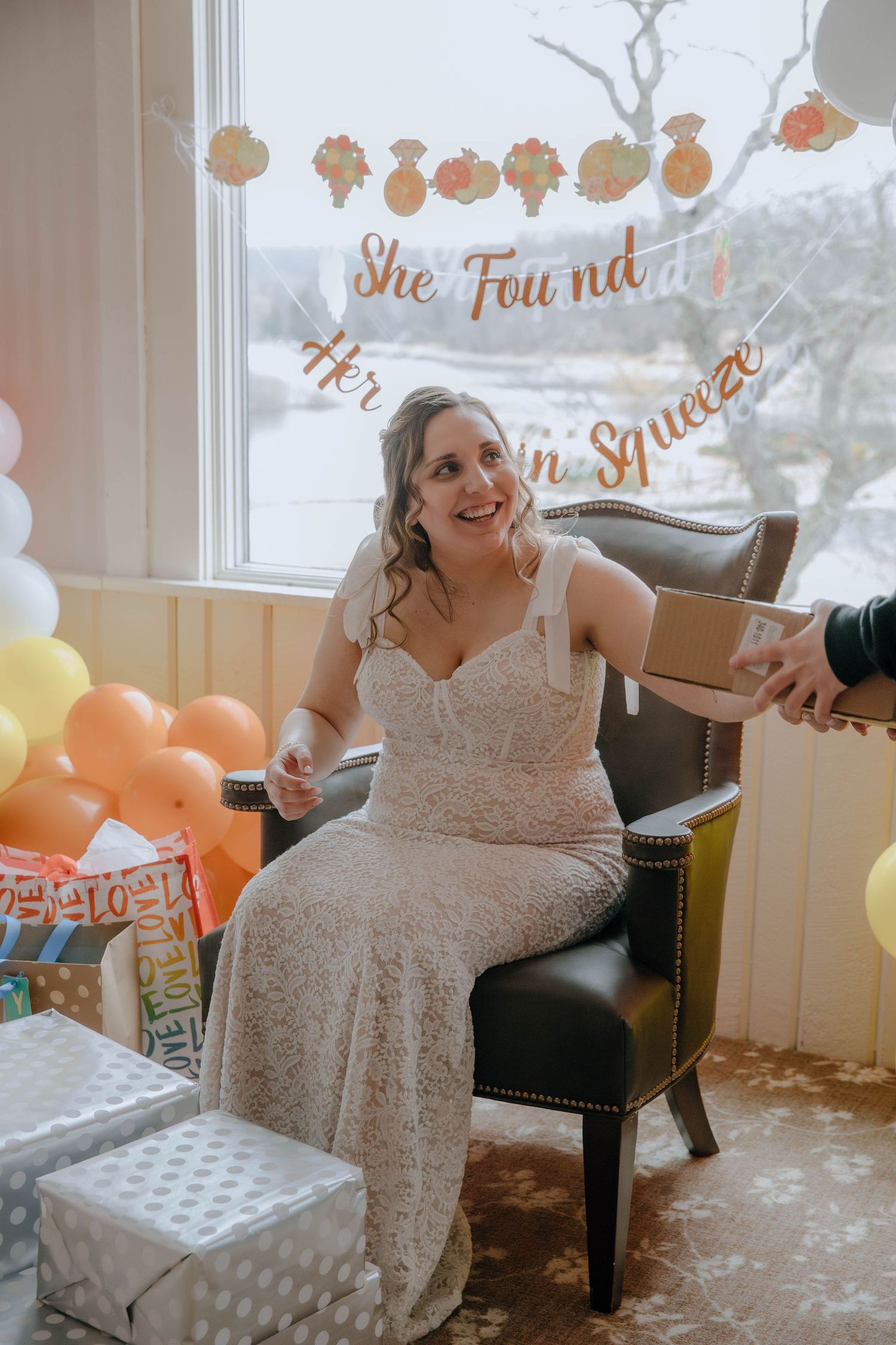 Bride in lace gown sits in chair at bridal shower, surrounded by gifts and balloons under a 