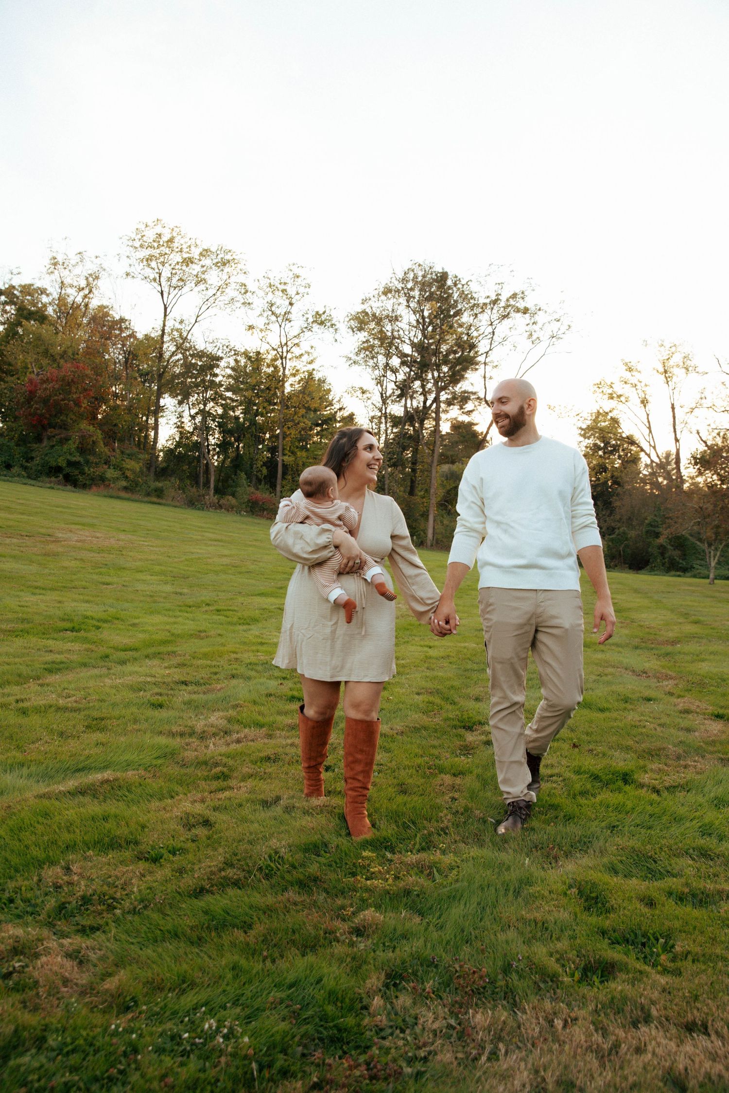 A young family walks hand-in-hand across a green field at sunset, with the mother holding their baby.