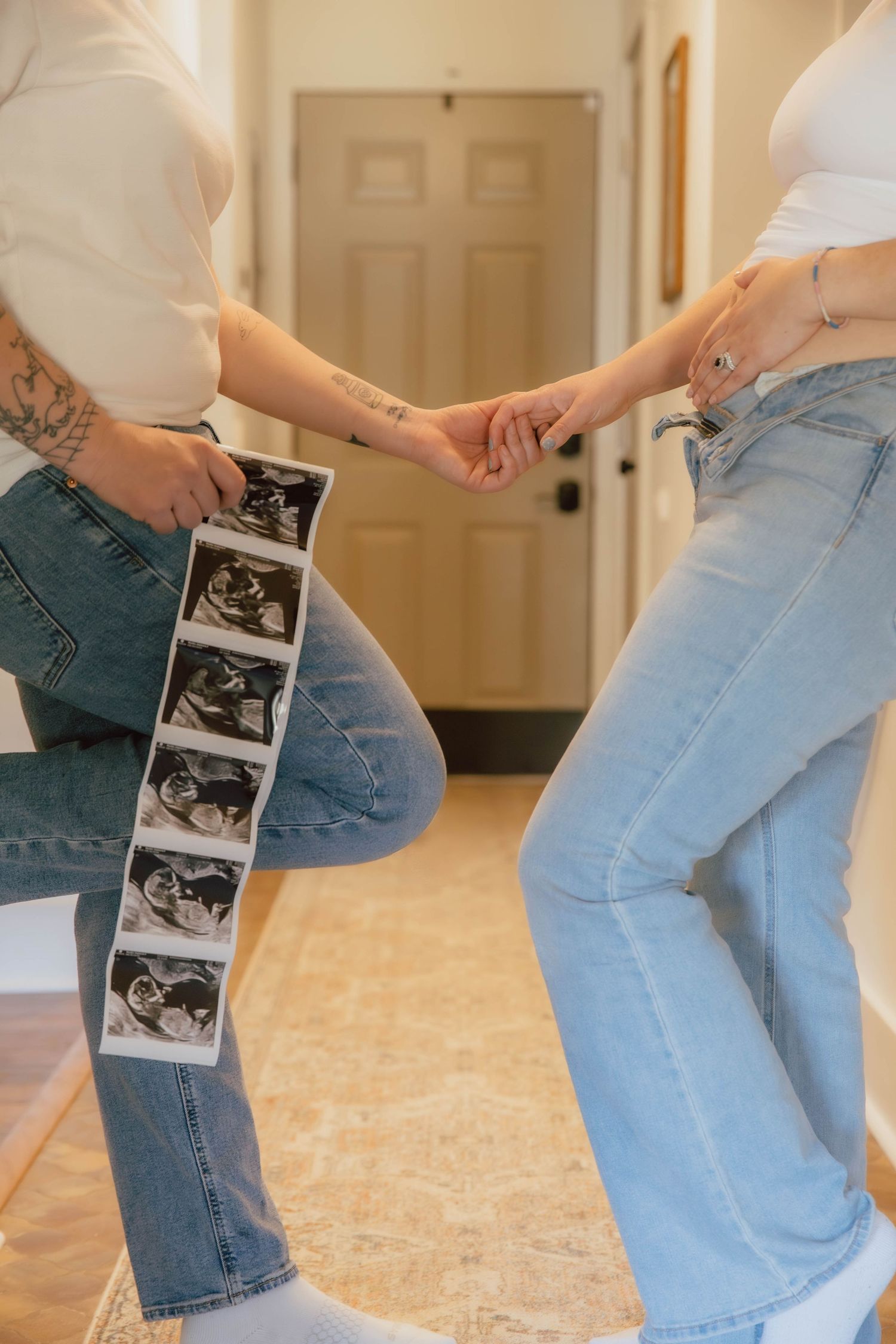 Two people holding hands while one displays a strip of ultrasound photos, wearing jeans indoors.