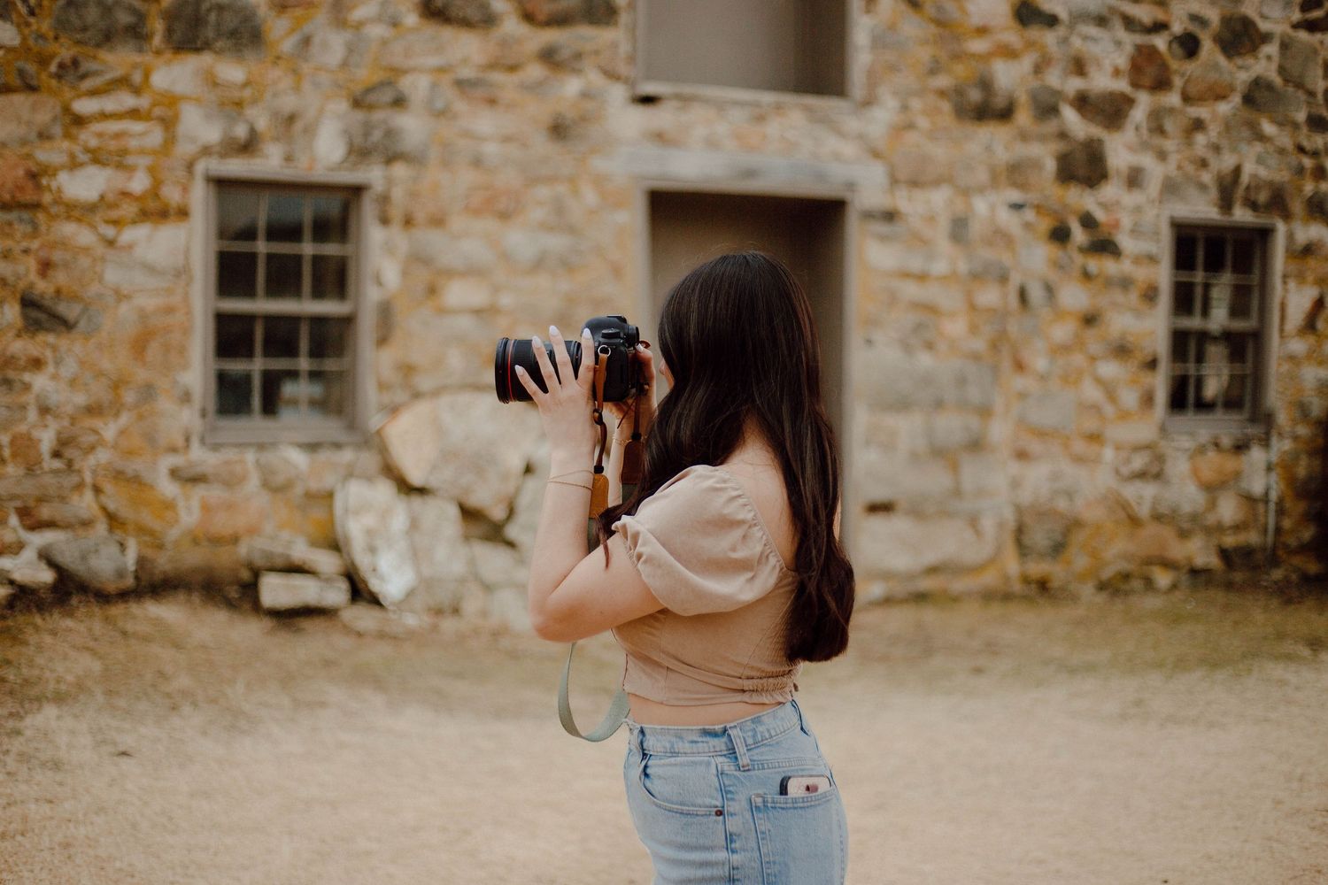 Young woman photographing rustic stone building with DSLR camera on sunny day outdoors.