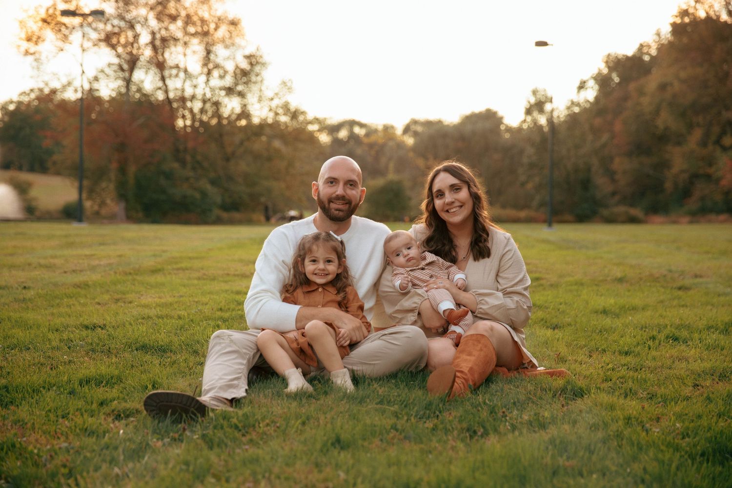 Smiling family of four posing together on lush green lawn surrounded by autumn trees at sunset.