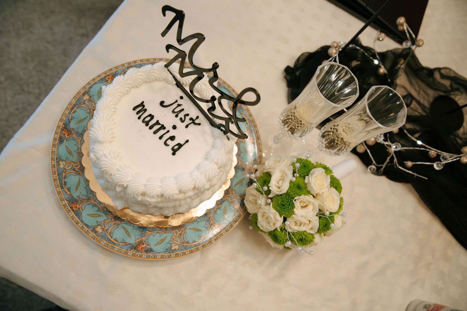 White two-tier just married wedding cake with Mr & Mrs topper, turquoise plate, and white rose bouquet on table.