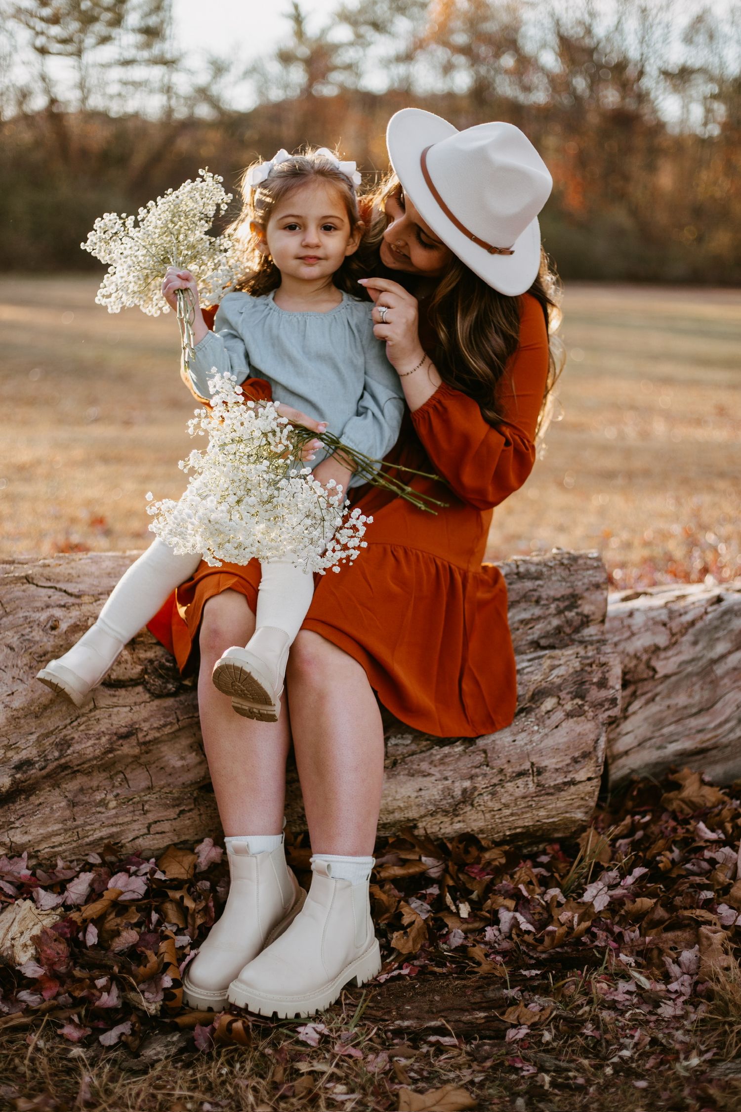 A mother in a rust dress and hat kisses her daughter holding white flowers on a log outdoors.
