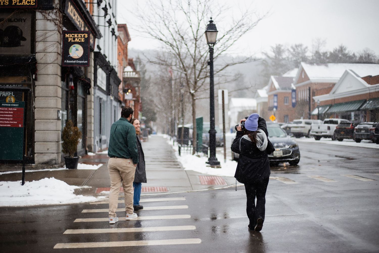 People walking on snowy small town sidewalk in winter with shops and street lamps visible.