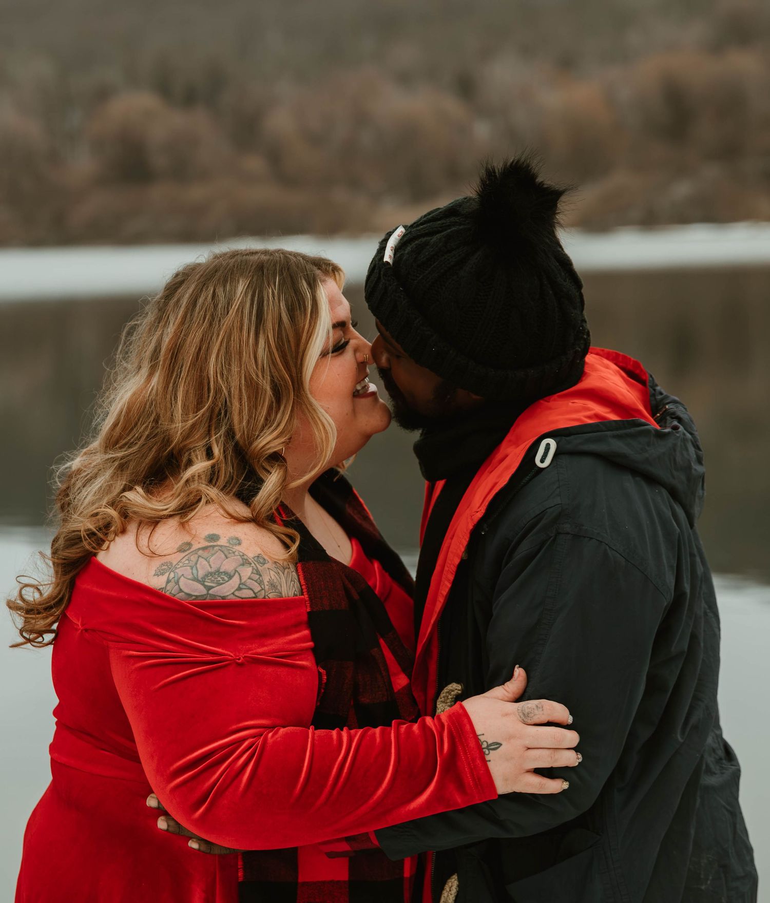 Couple sharing a romantic kiss outdoors in winter, woman in red velvet dress with shoulder tattoo, man in black jacket.