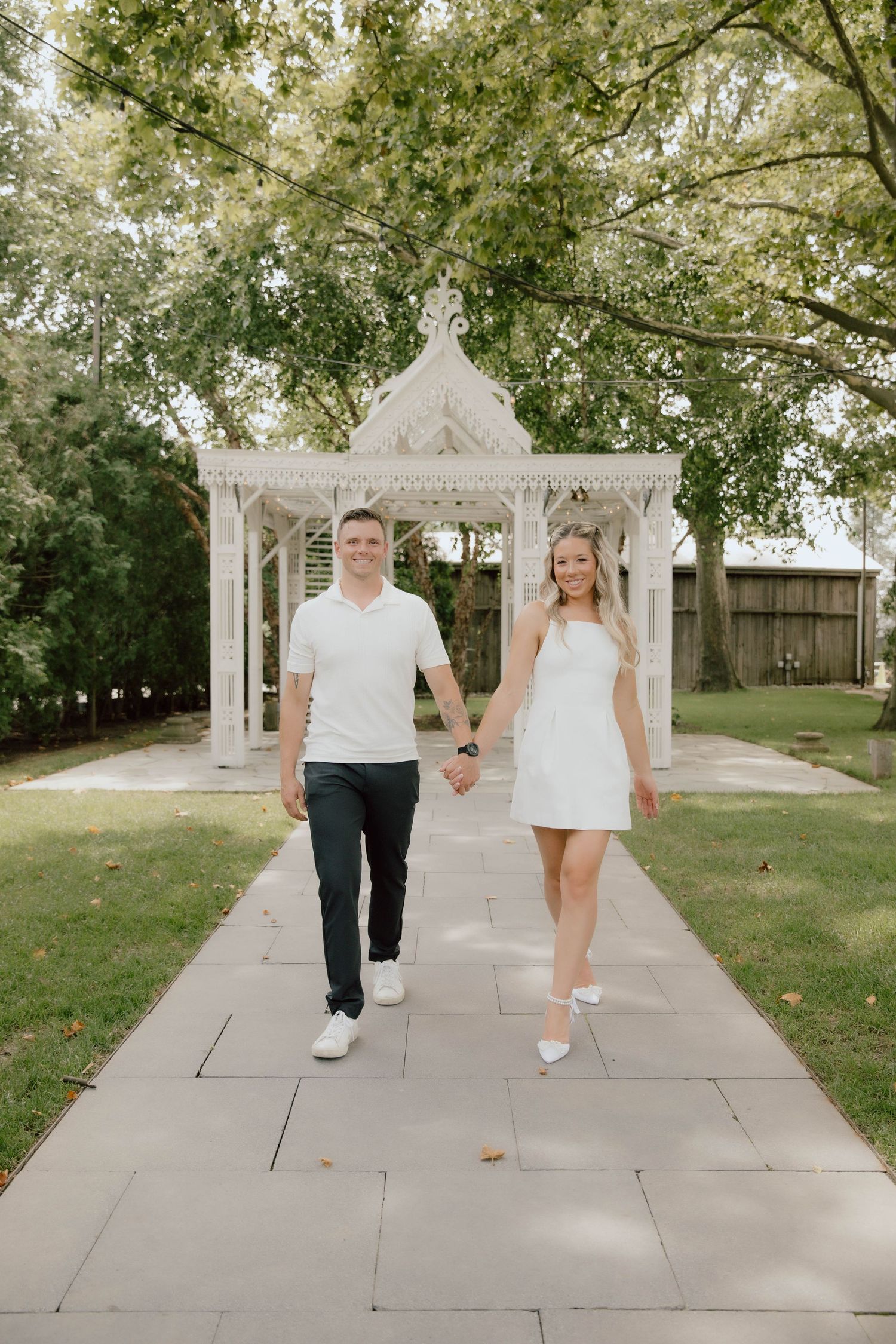 Smiling couple holding hands and walking toward camera on a garden path with white gazebo behind them.
