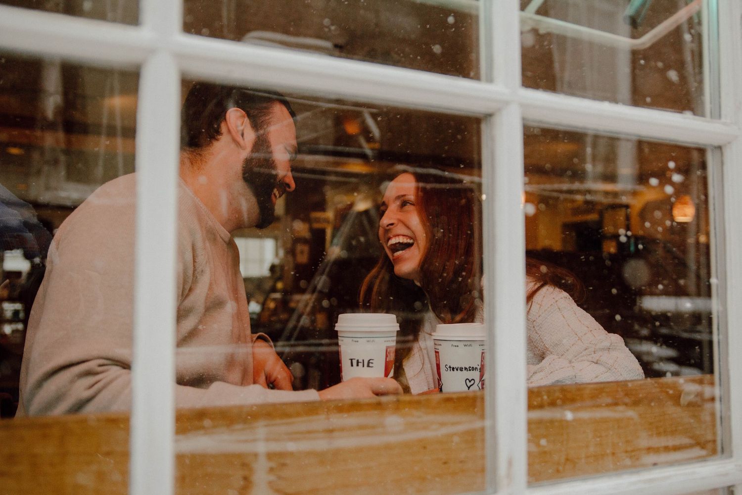 Happy couple laughing together over coffee cups inside cozy cafe viewed through snow-dusted window.