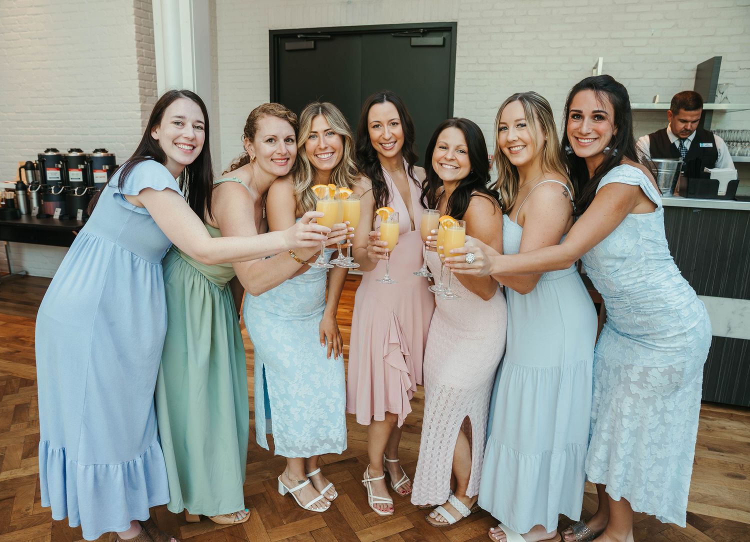 Seven women in pastel dresses toasting with drinks at a wedding reception celebration.