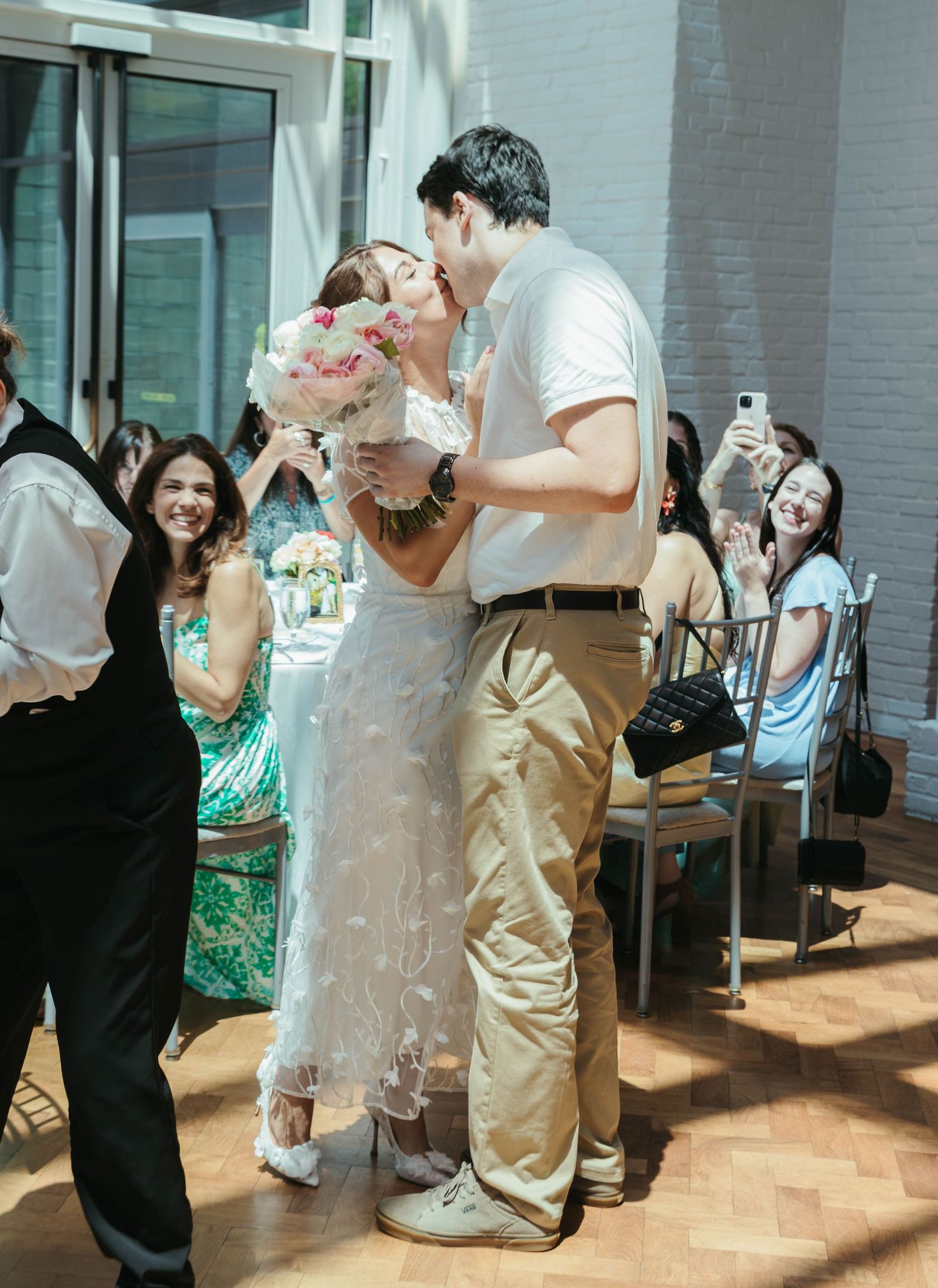 A bride and groom share a kiss at their wedding reception while guests smile and celebrate around them.