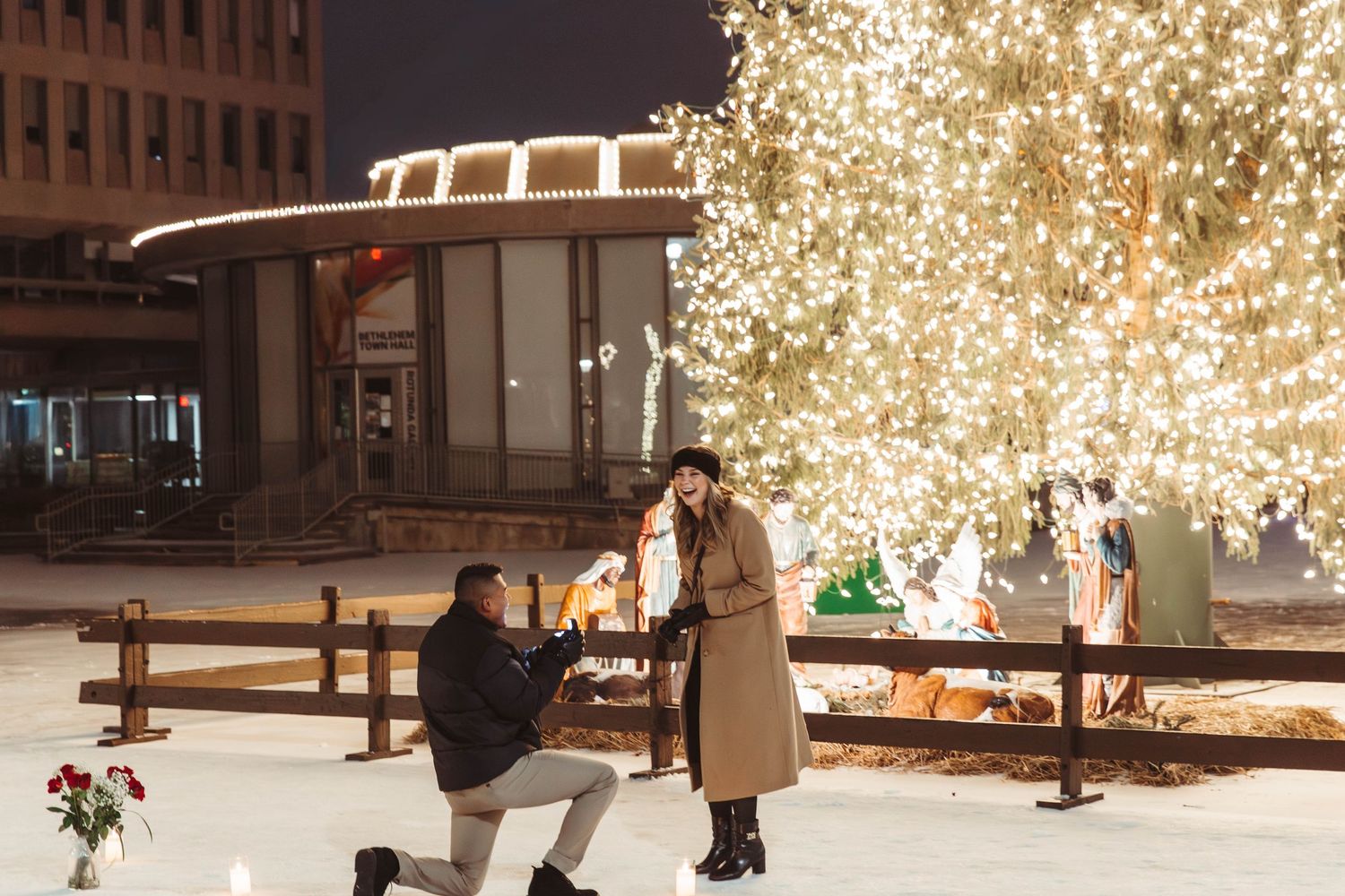 Man proposing to woman on one knee near a glowing Christmas tree at night outdoors.
