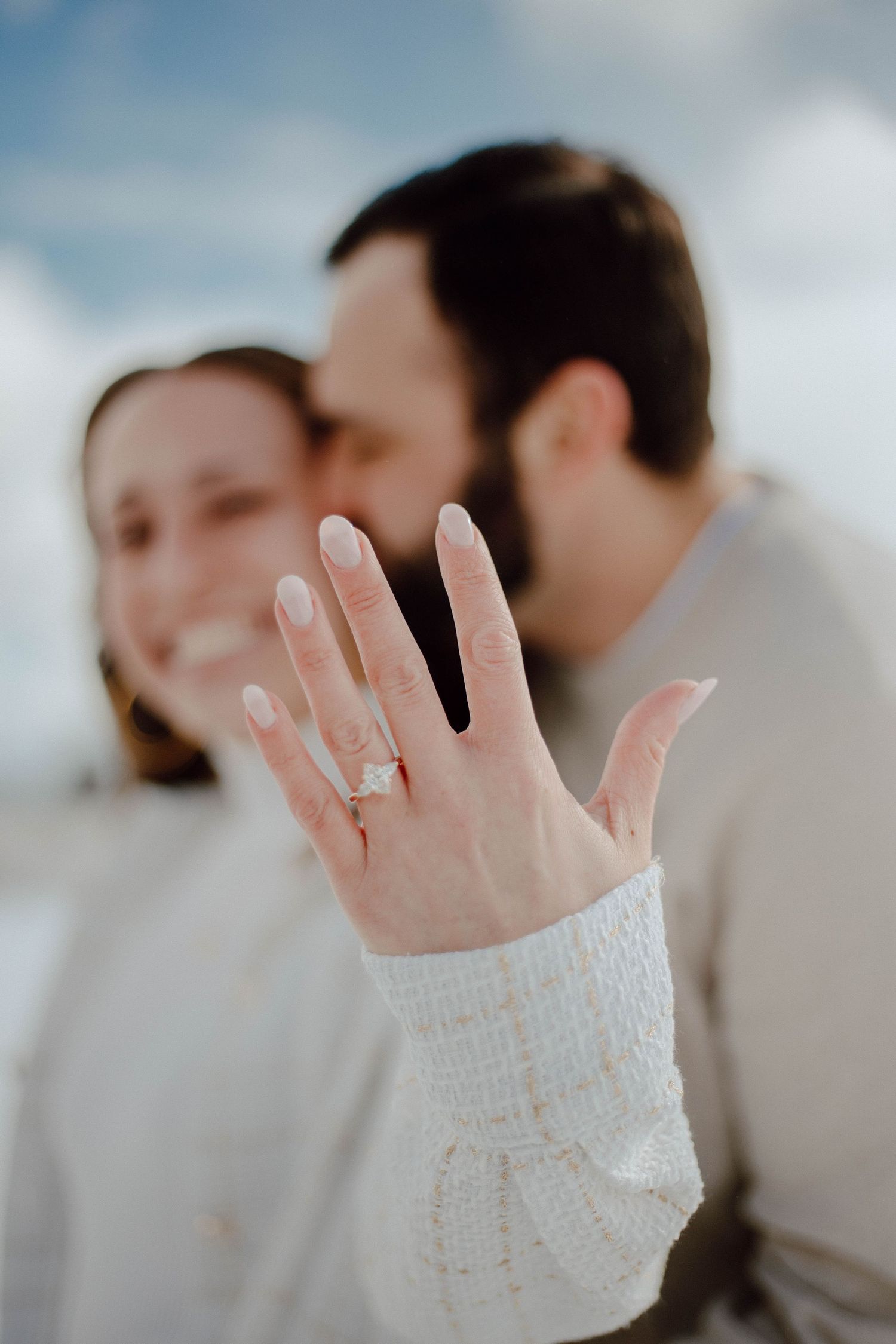 Engaged couple showing off diamond ring in foreground while sharing a kiss in soft focus background.