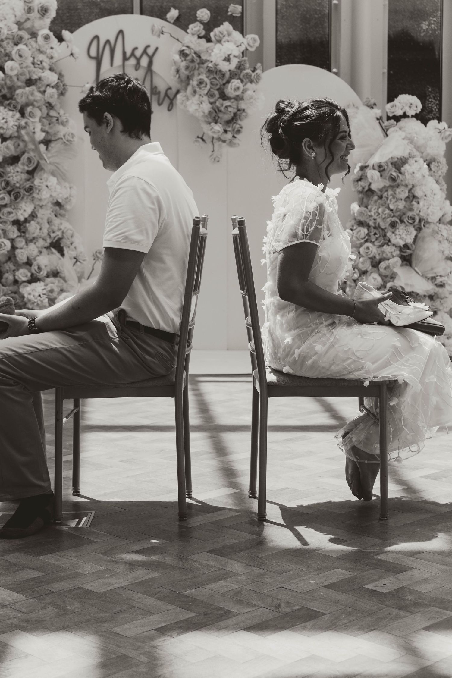 Black and white photo of bride and groom sitting back-to-back on chairs at wedding venue with floral backdrop.