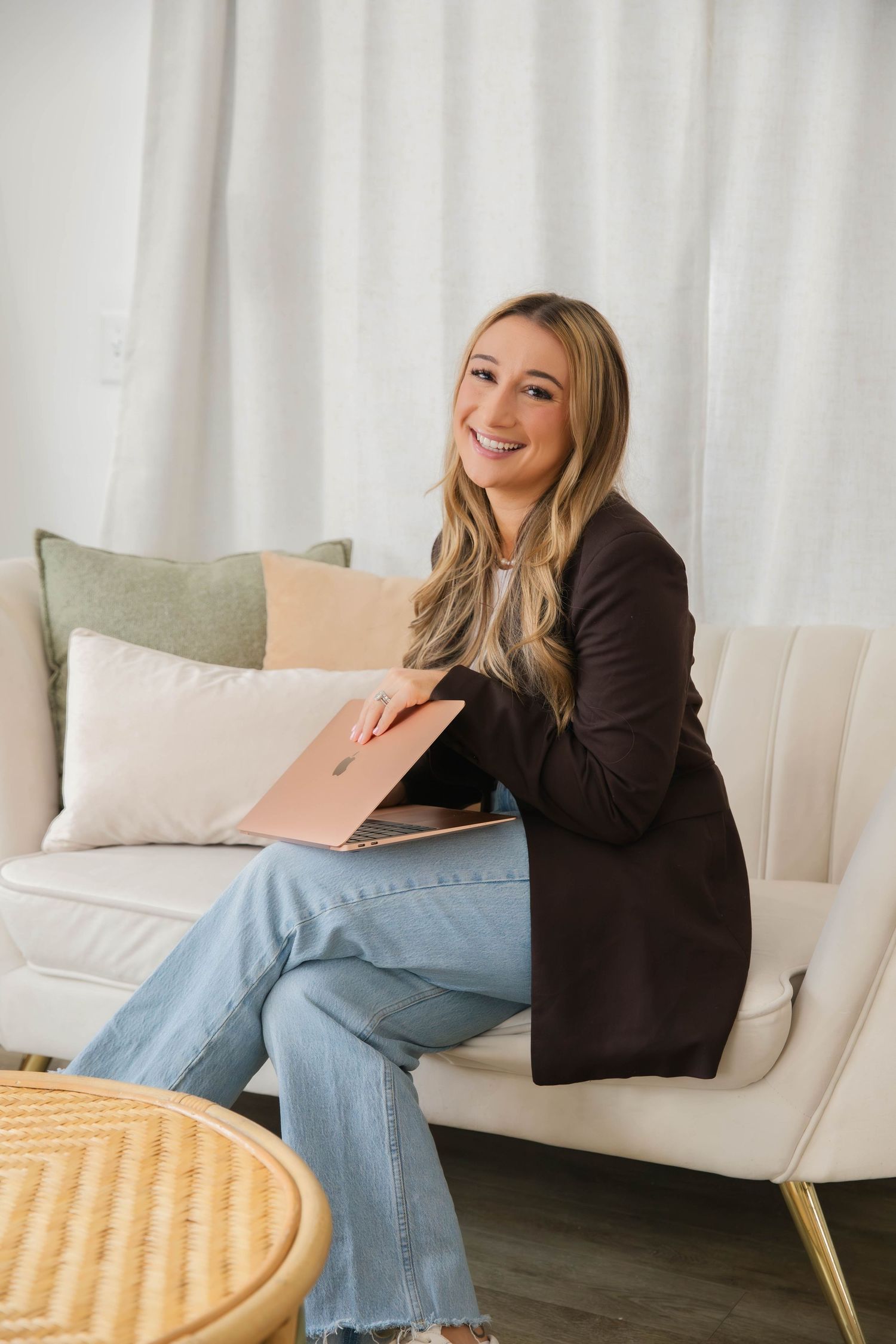 Smiling blonde woman in dark blazer and jeans sitting on white sofa holding rose gold laptop.