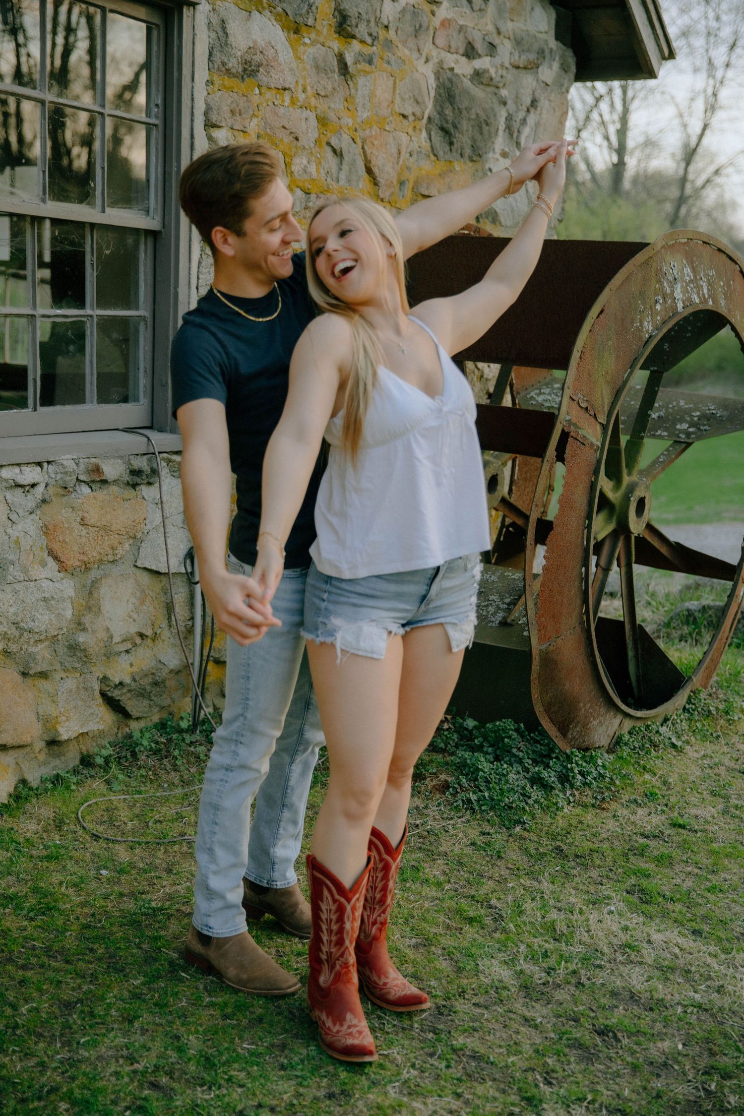 A smiling couple poses near a rustic stone building with a vintage wagon wheel, woman wearing red cowboy boots.
