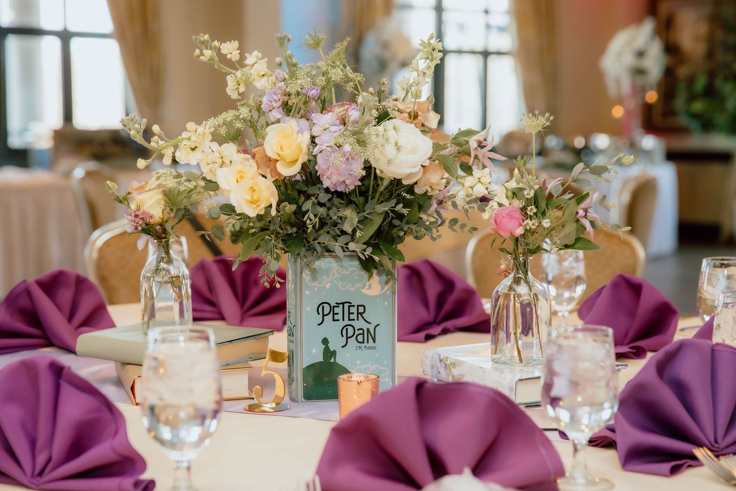 Elegant wedding reception table with purple napkins, floral centerpiece, and a Peter Pan themed table number card.