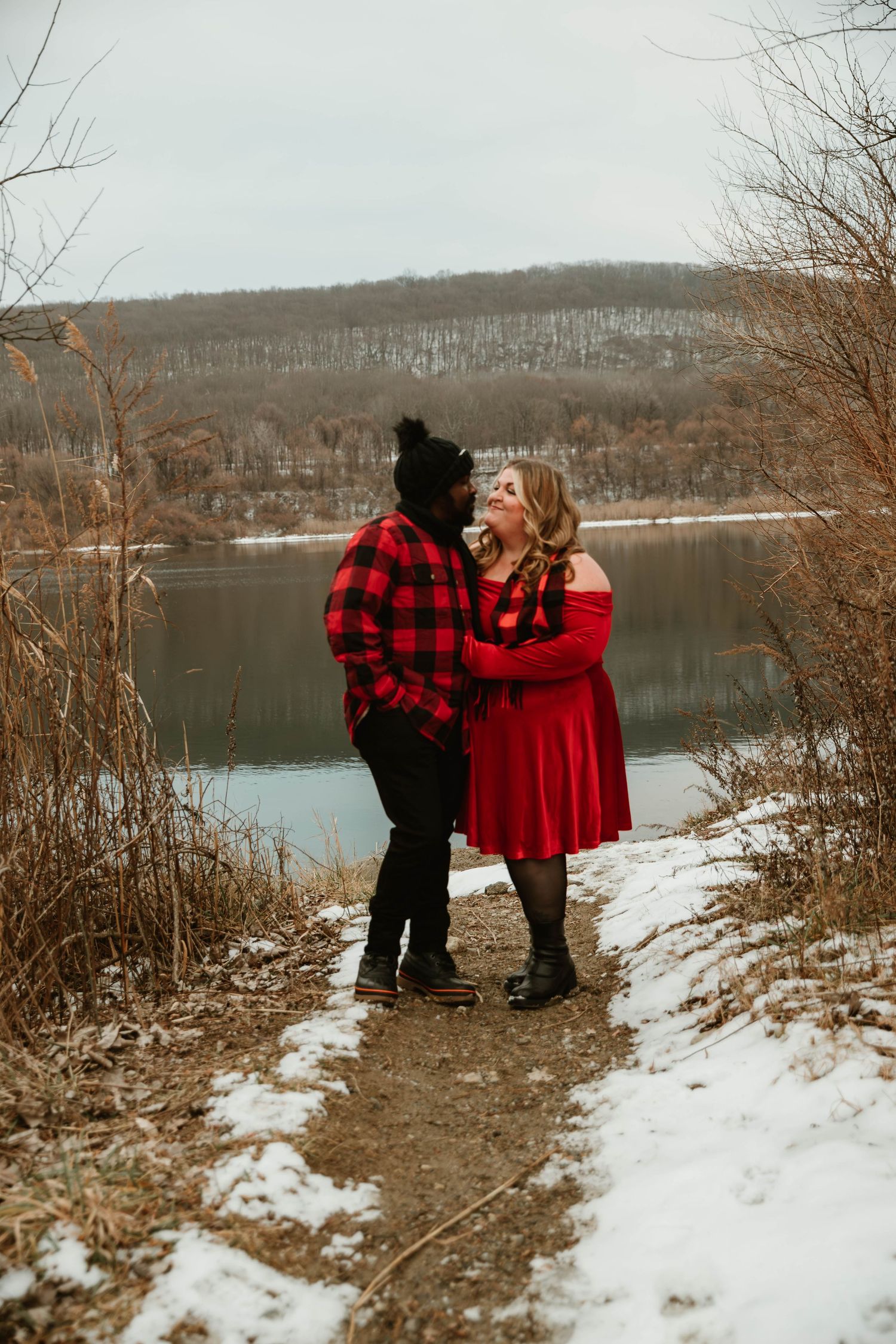 Couple in red outfits sharing a romantic moment on a snowy lakeside trail in winter.