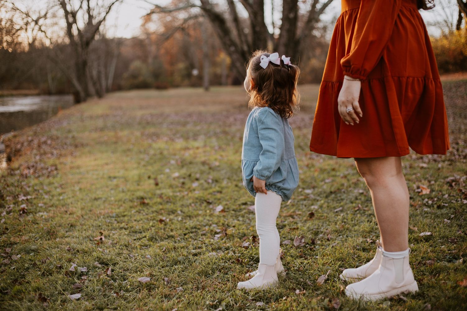 A young girl in a blue outfit and white boots stands beside a woman in a rust dress outdoors in a park.