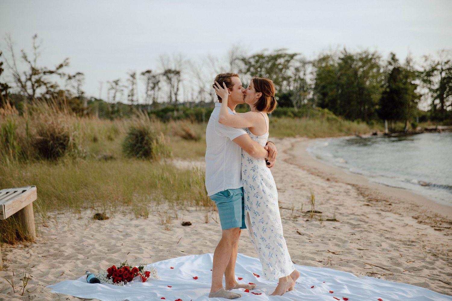 Couple sharing a romantic kiss on a sandy beach with rose petals and a white blanket during a marriage proposal.