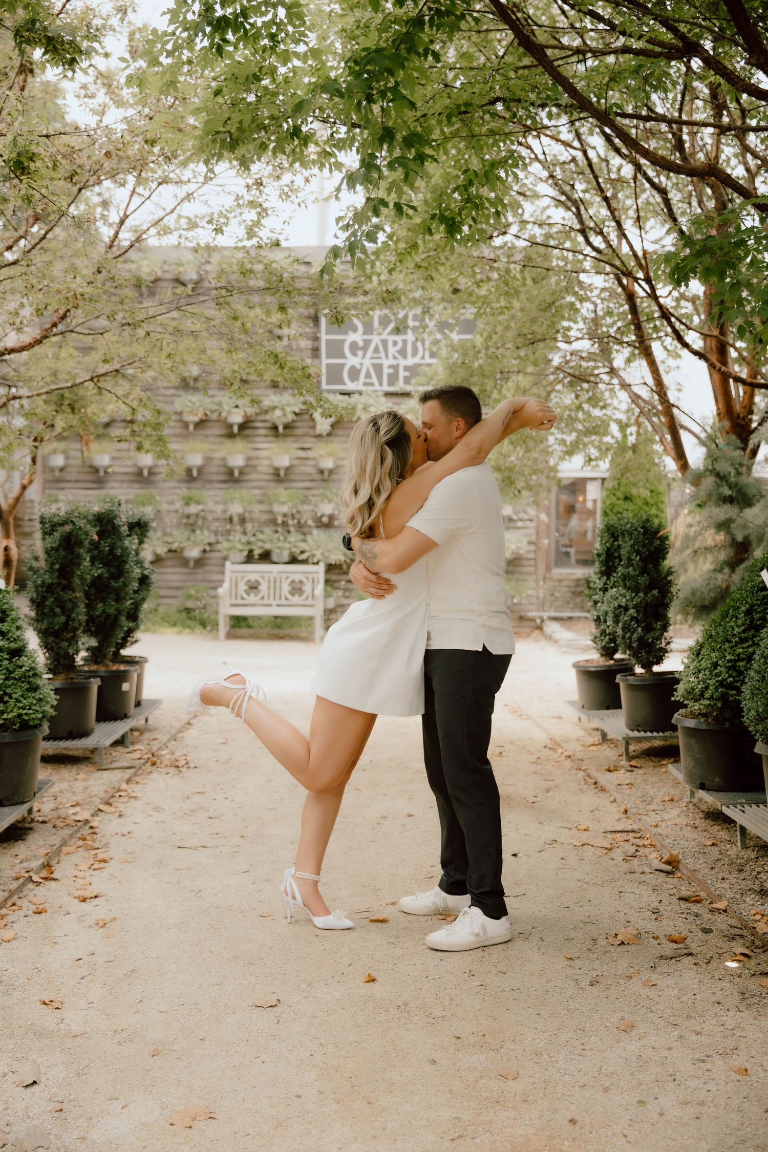 Couple embracing and kissing on a garden path near a cafe, surrounded by lush trees and potted topiaries.