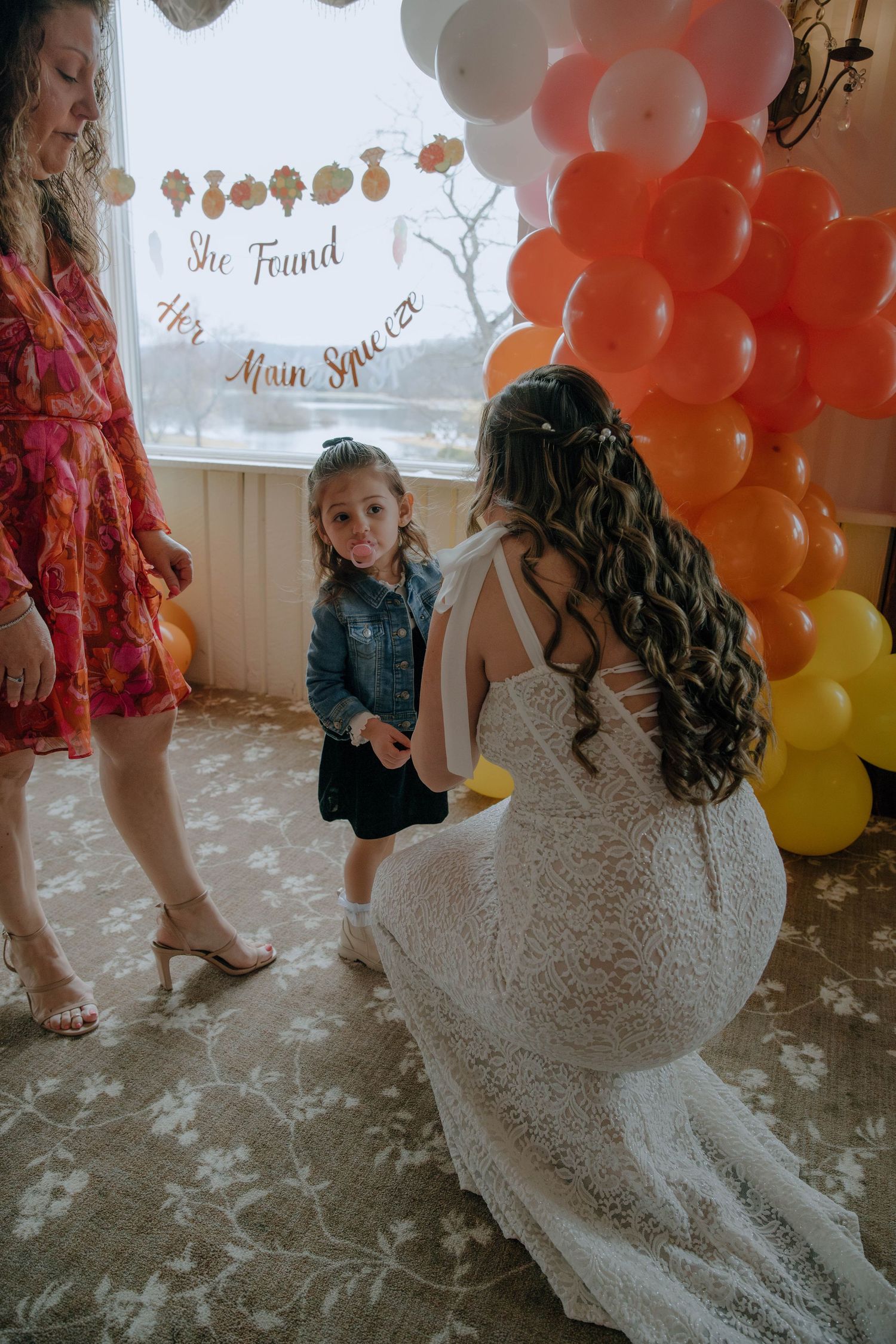 Bride in lace gown kneeling to greet young girl near colorful balloon arch at bridal shower.