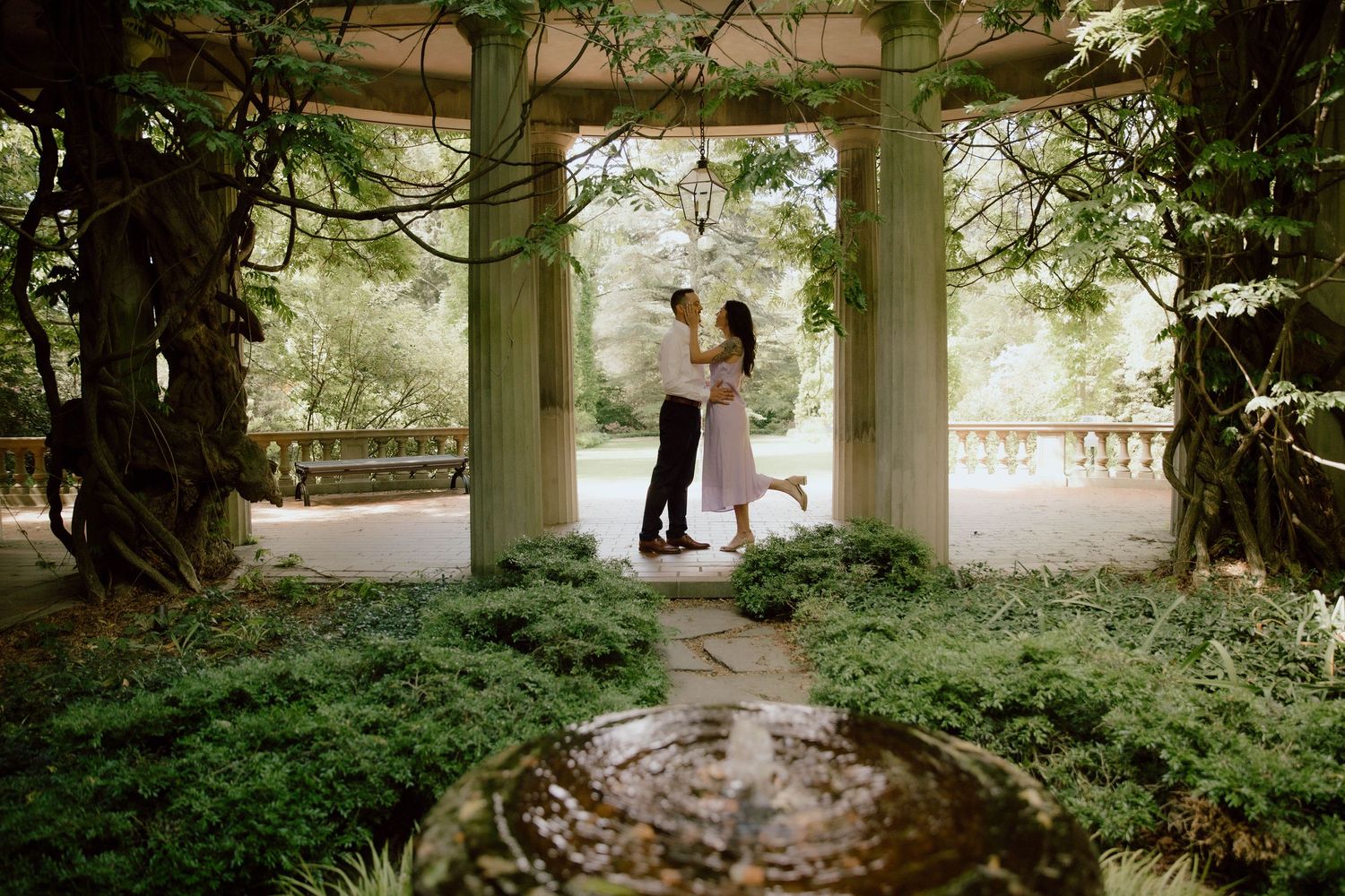 Romantic couple embracing under a garden gazebo surrounded by lush green foliage and a stone fountain.