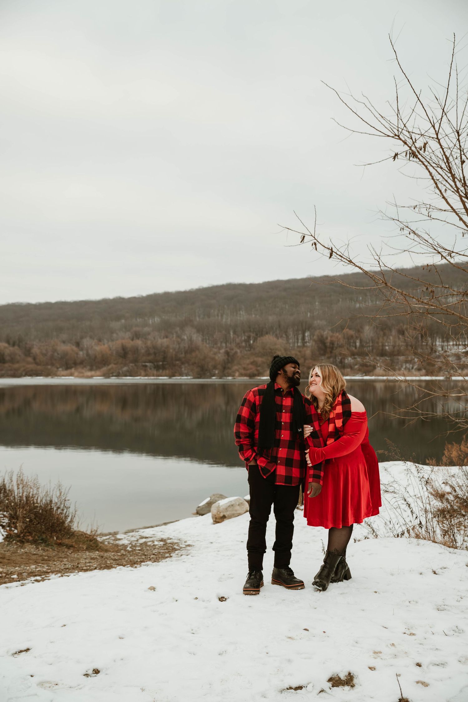 Couple in matching red outfits sharing a romantic moment by a snowy lakeside during a winter engagement photo session.