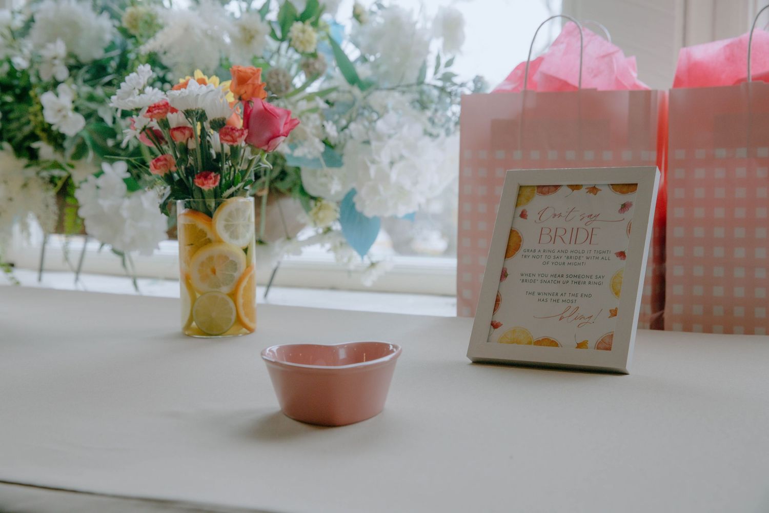 Elegant bridal shower table with pink bowl, framed invite, gift bags, and floral arrangements near a bright window.