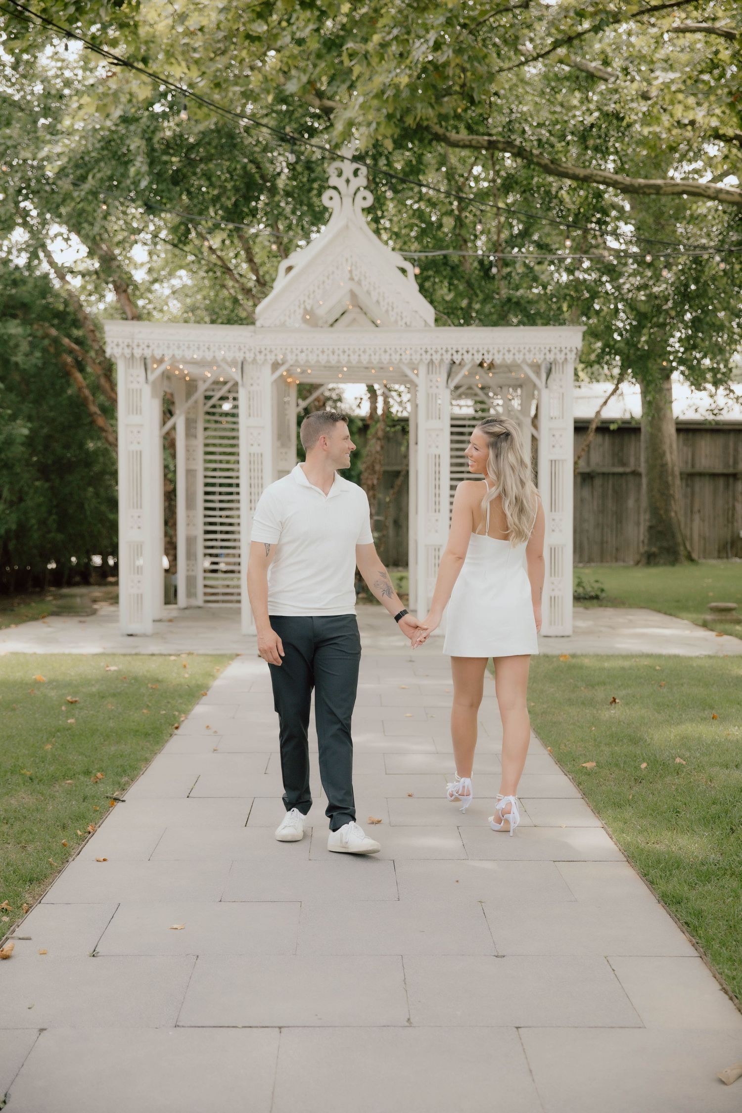 Couple in matching white outfits holding hands on a garden path with a white ornate gazebo backdrop.