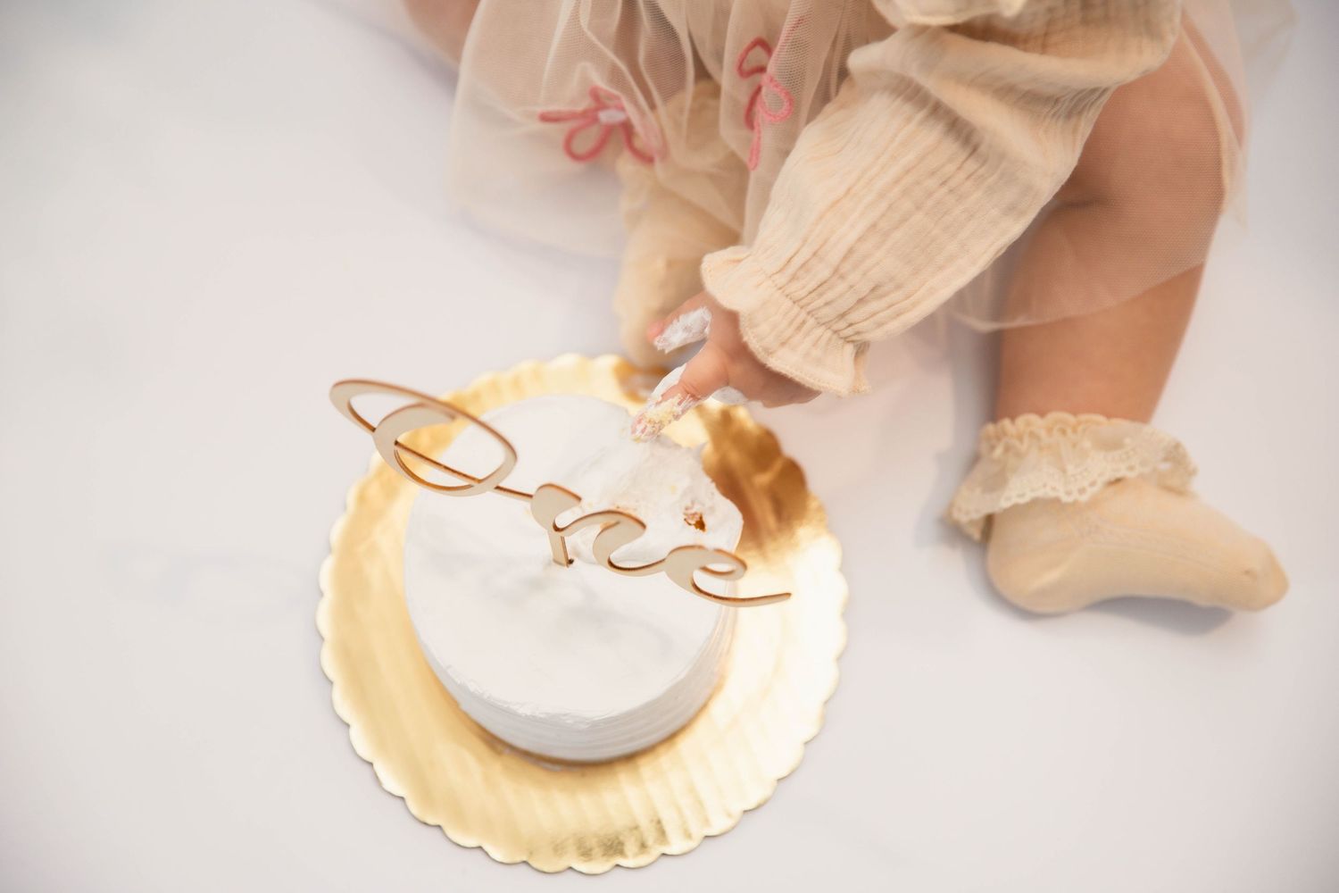 Baby girl in cream tutu smashing a white and gold first birthday cake with \