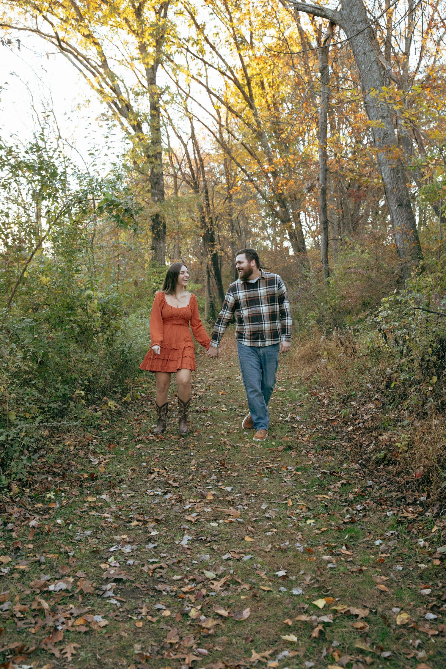 Couple holding hands walking on a leaf-covered forest path in autumn, woman in orange dress and man in plaid shirt.