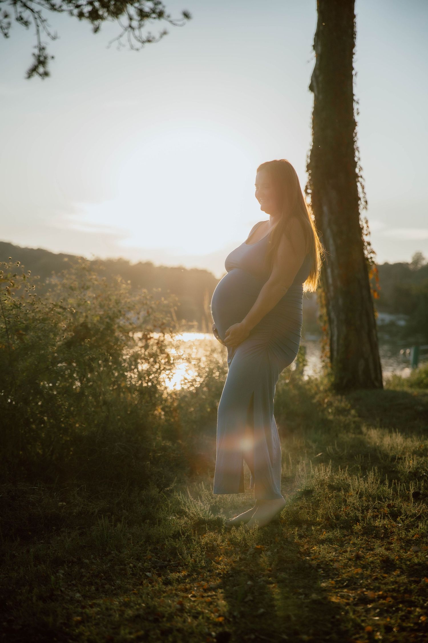 Pregnant woman in gray dress holding belly at golden hour sunset outdoors near trees.