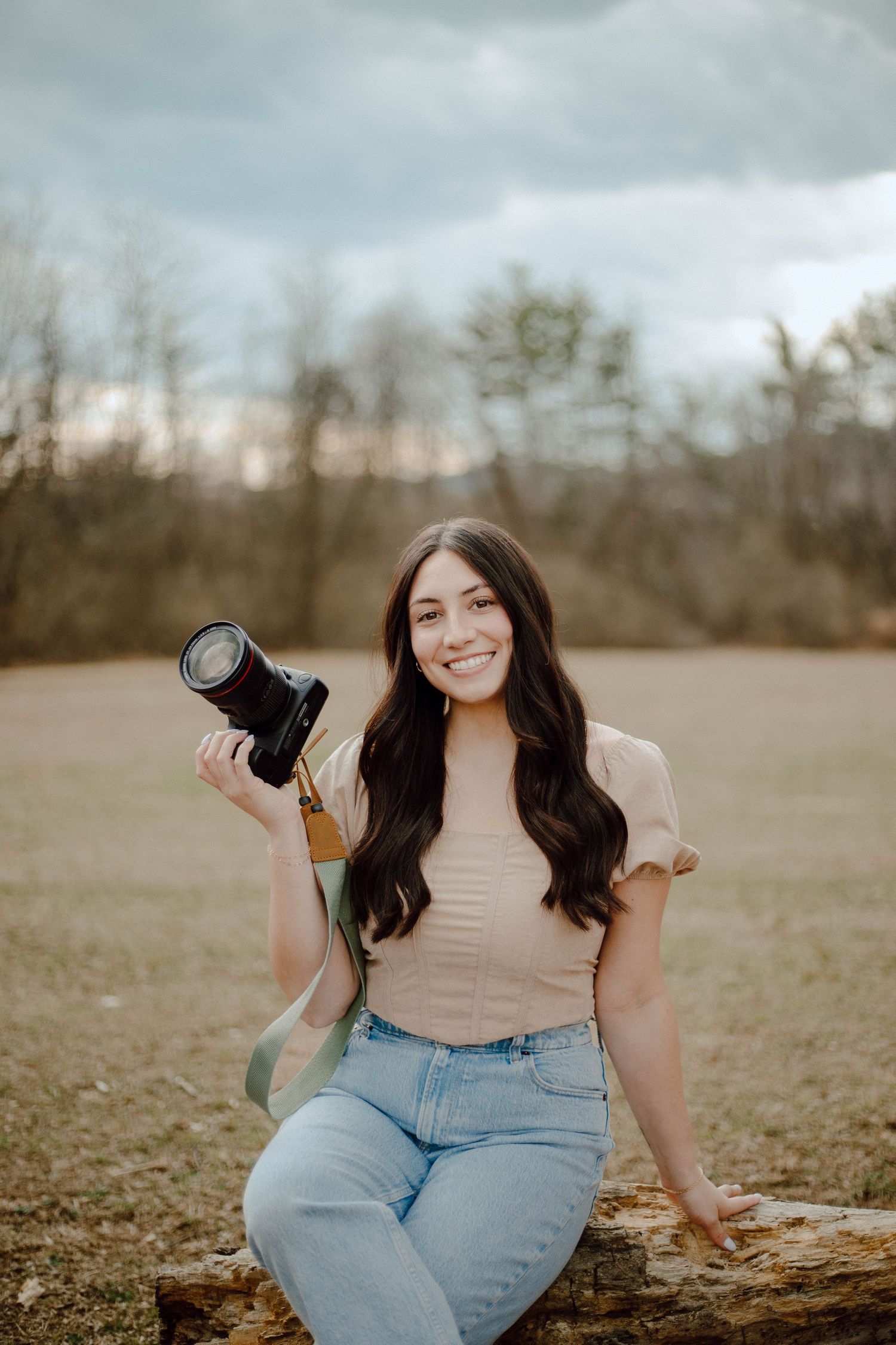 Young woman smiling outdoors holding a DSLR camera, wearing jeans and a beige top, seated on a log in a field.