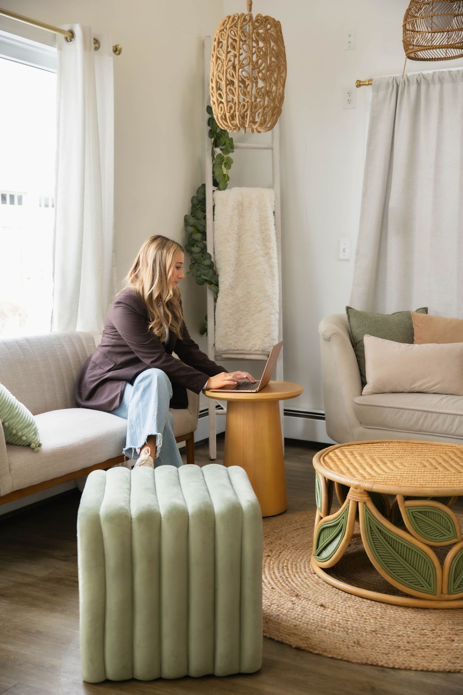 Blonde woman working on laptop in bright boho-style living room with rattan furniture.