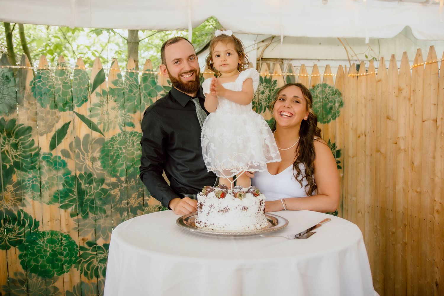 A couple and young child celebrate at a wedding cake table outdoors, with a white fence and tropical backdrop.
