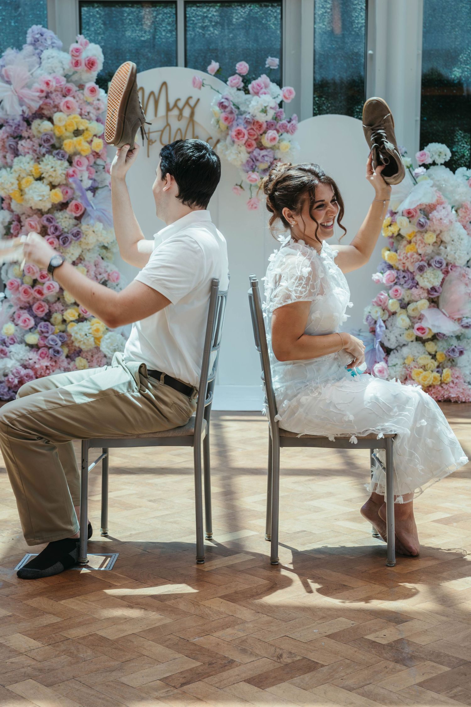Bride and groom playing the shoe game at their wedding reception in front of a colorful floral backdrop.