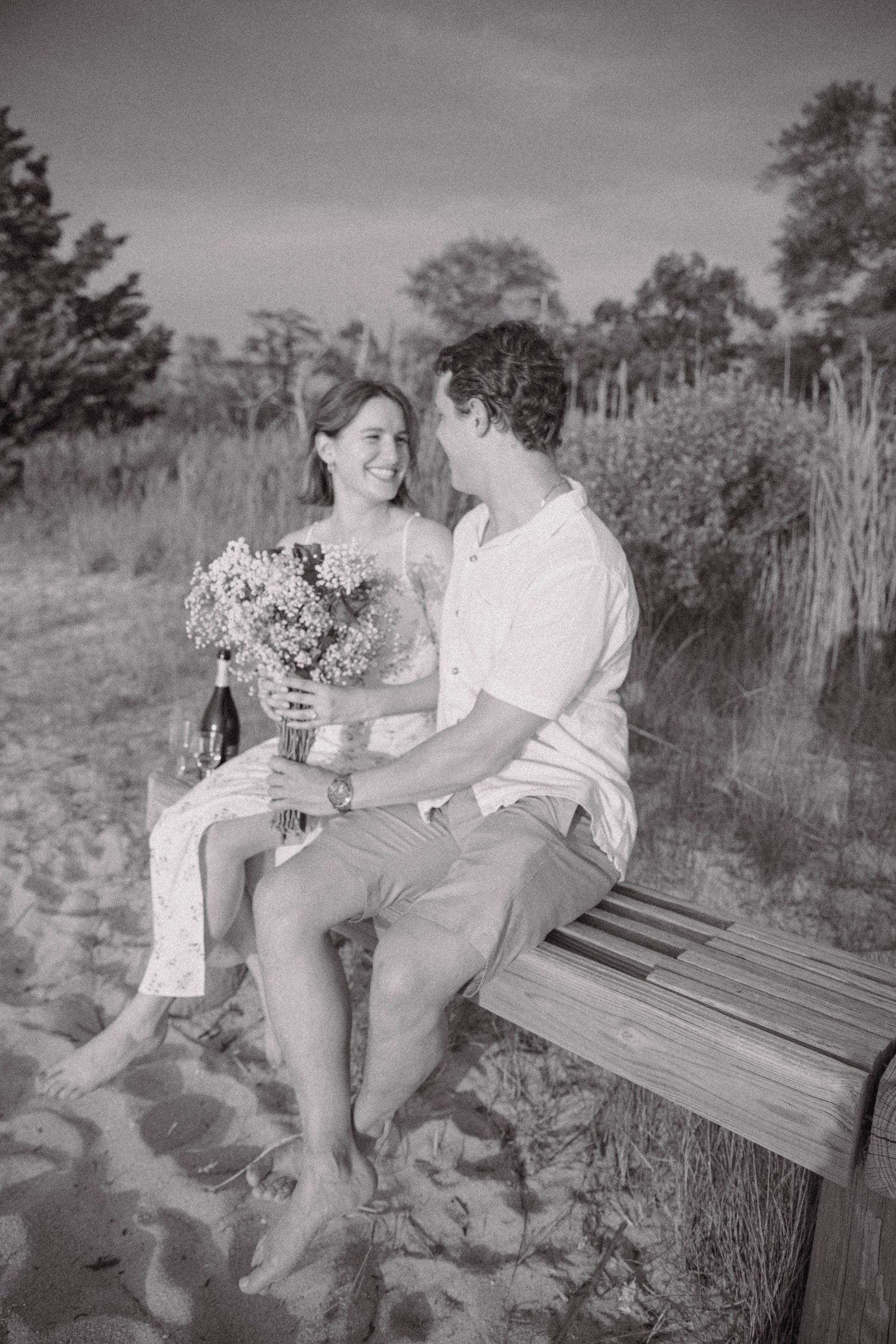 Black and white photo of a couple sitting on a bench at the beach with flowers.