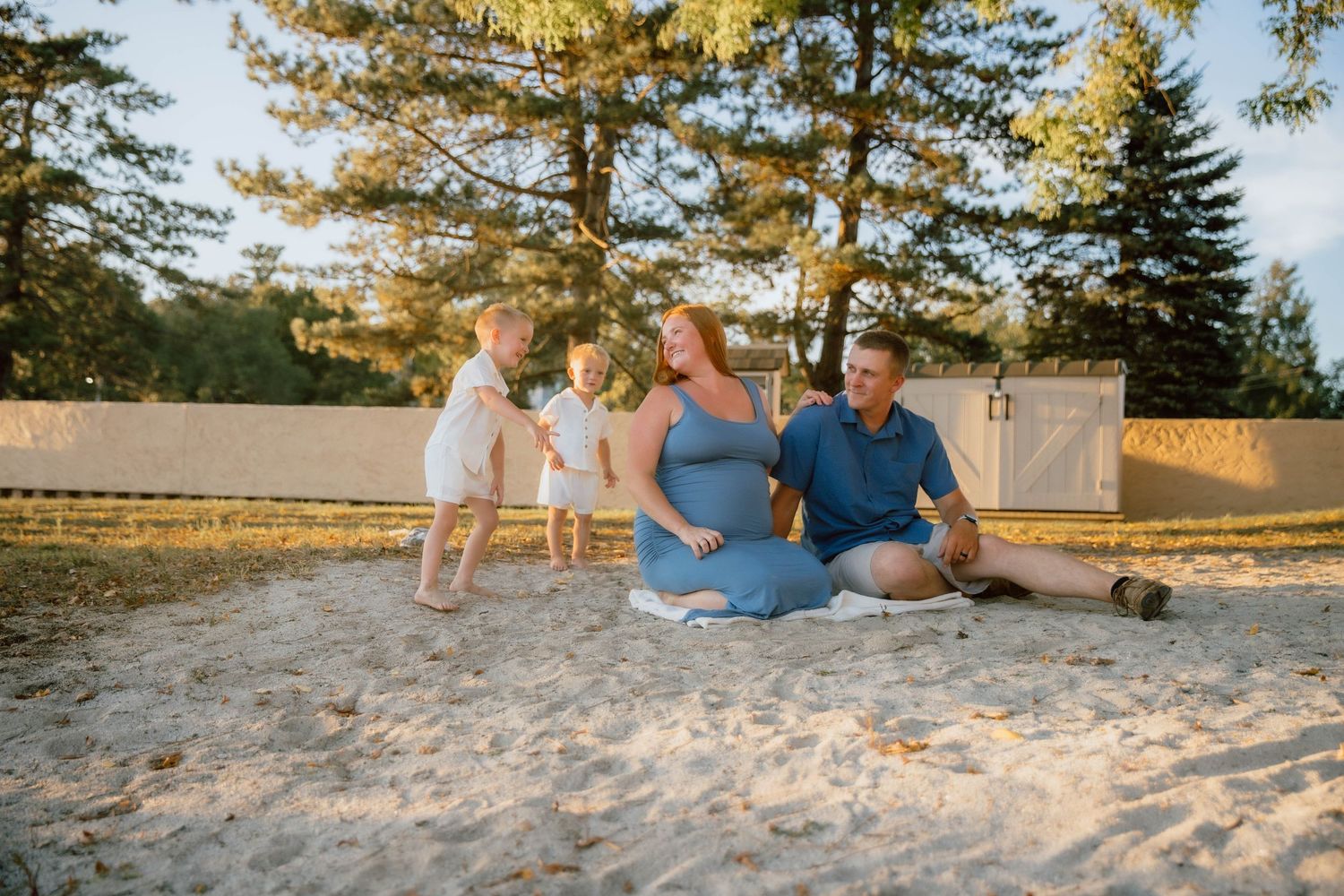 A happy family with two toddlers sits together on sandy ground at golden hour.