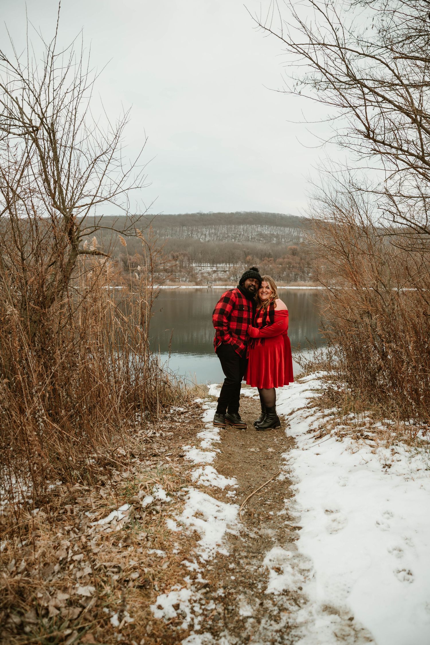 Couple in red outfits embracing on snowy lakeside trail surrounded by bare winter trees.