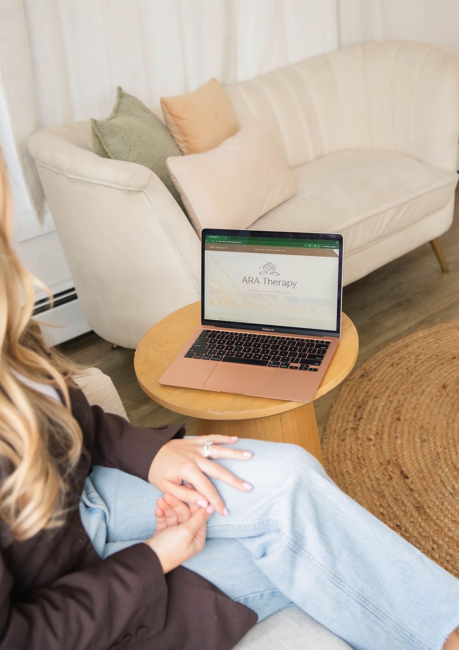 Woman sitting with laptop on round table displaying ARA Therapy website in a cozy living room setting.