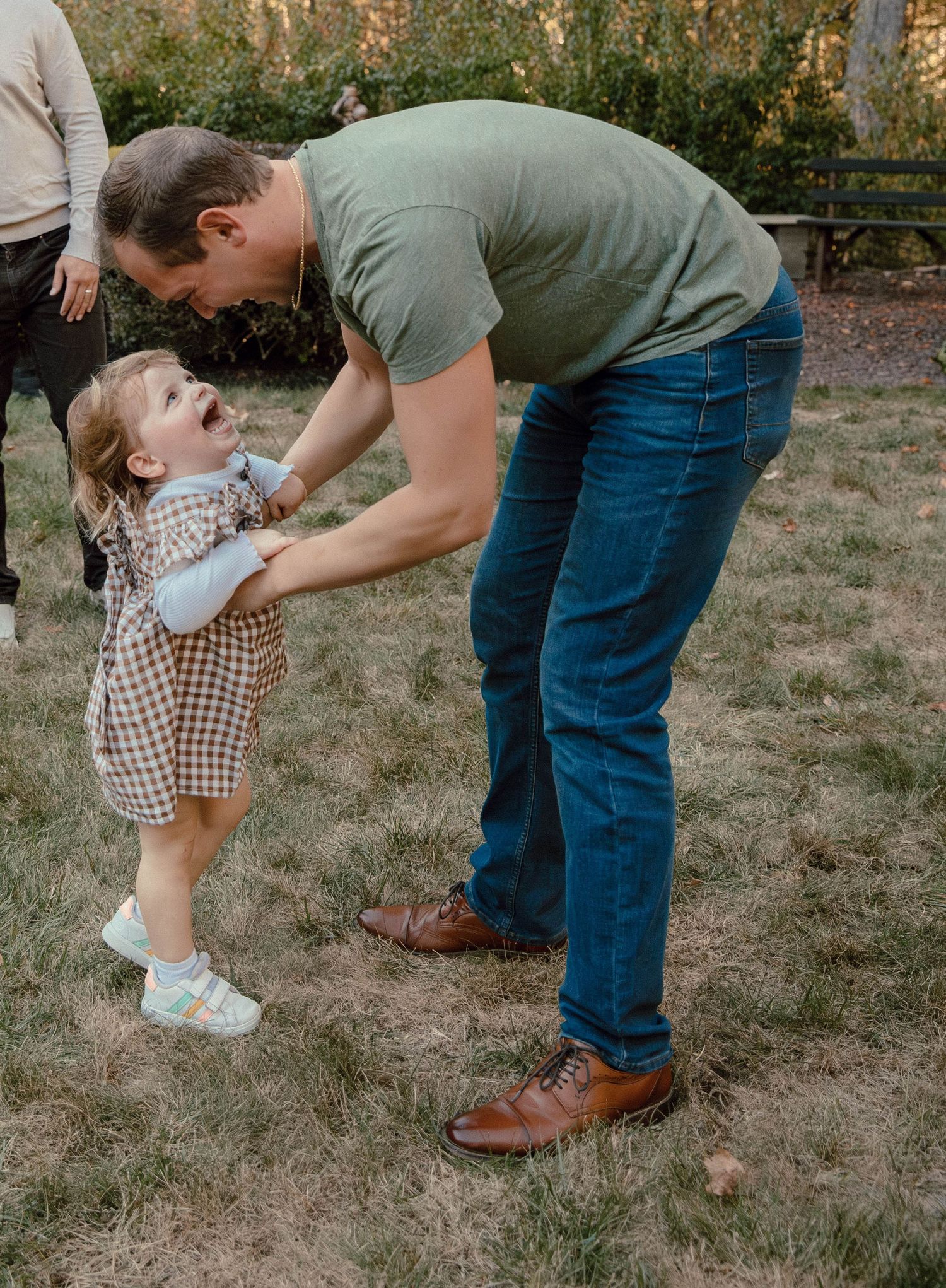 A man in a green shirt and jeans bends down to hold a laughing toddler girl in a plaid dress outdoors.
