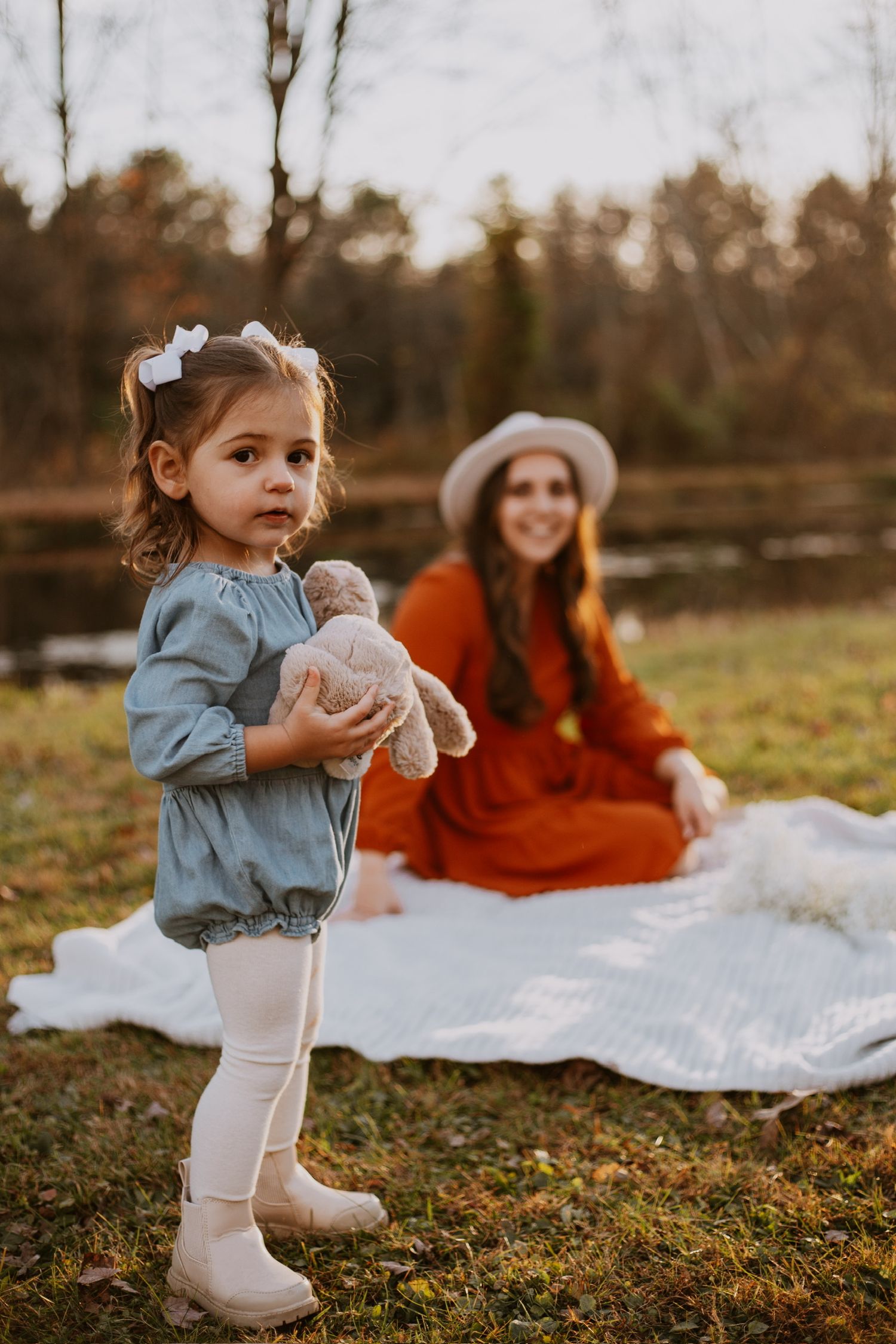 A toddler girl in a blue dress holds a stuffed bunny outdoors while her mother in an orange dress sits on a blanket.