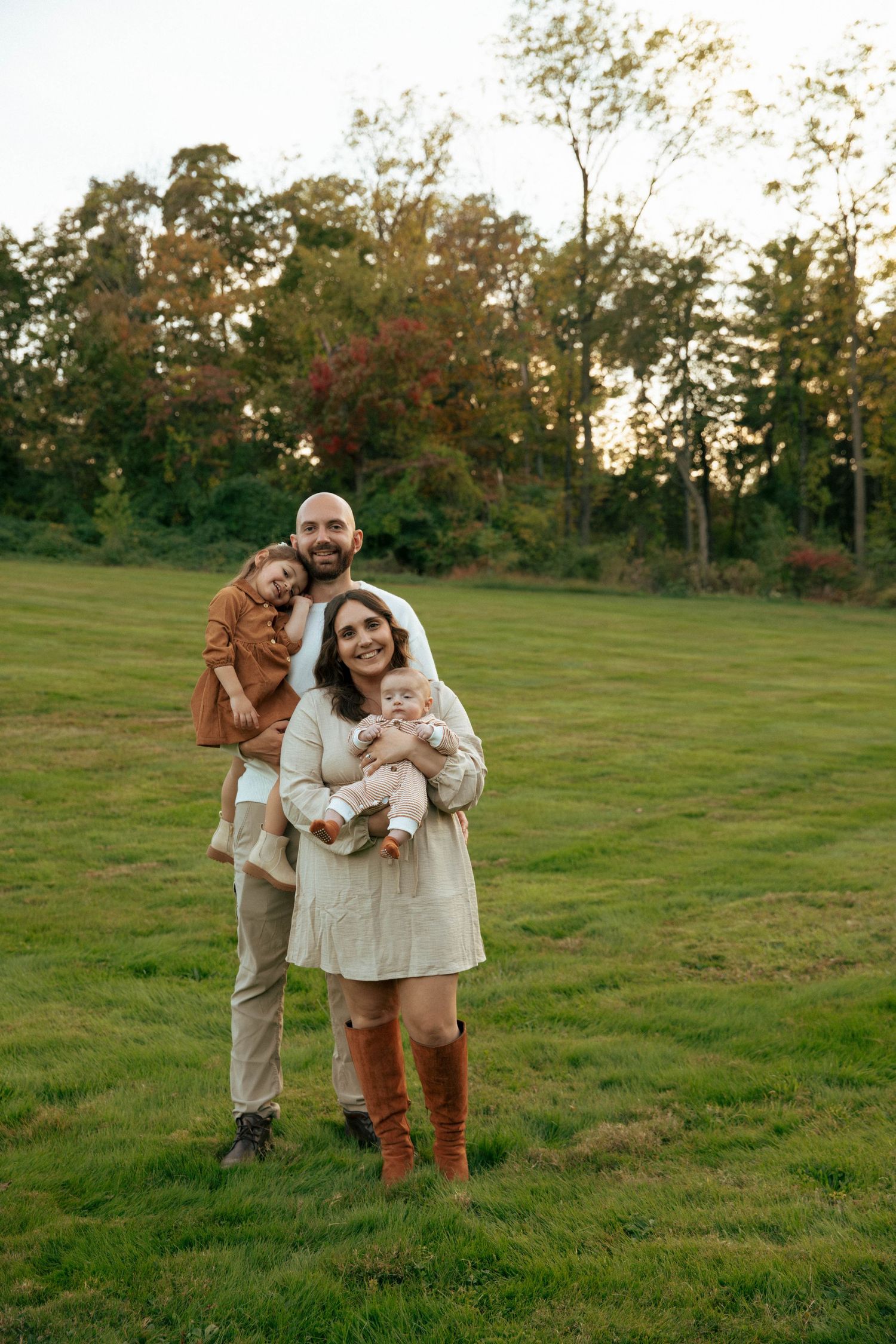 Family of four posing outdoors on green grass field at sunset, parents holding two young children, surrounded by autumn trees.