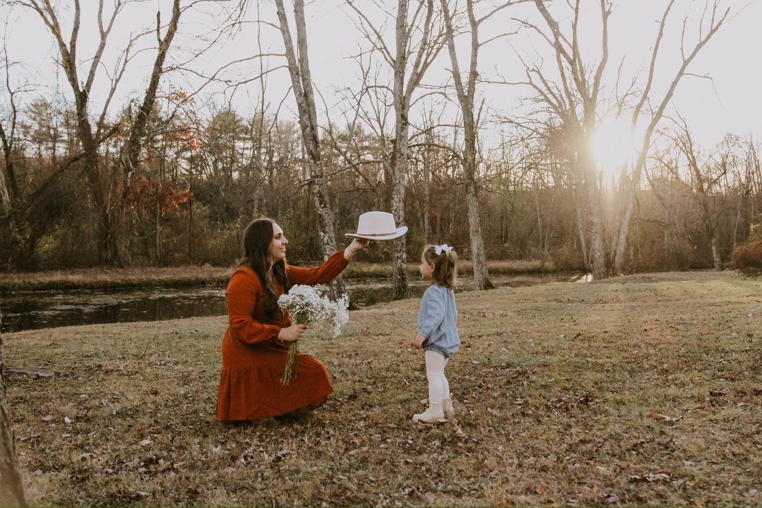 A mother in an orange dress holds a white hat for her young daughter outdoors in a sunlit autumn field.